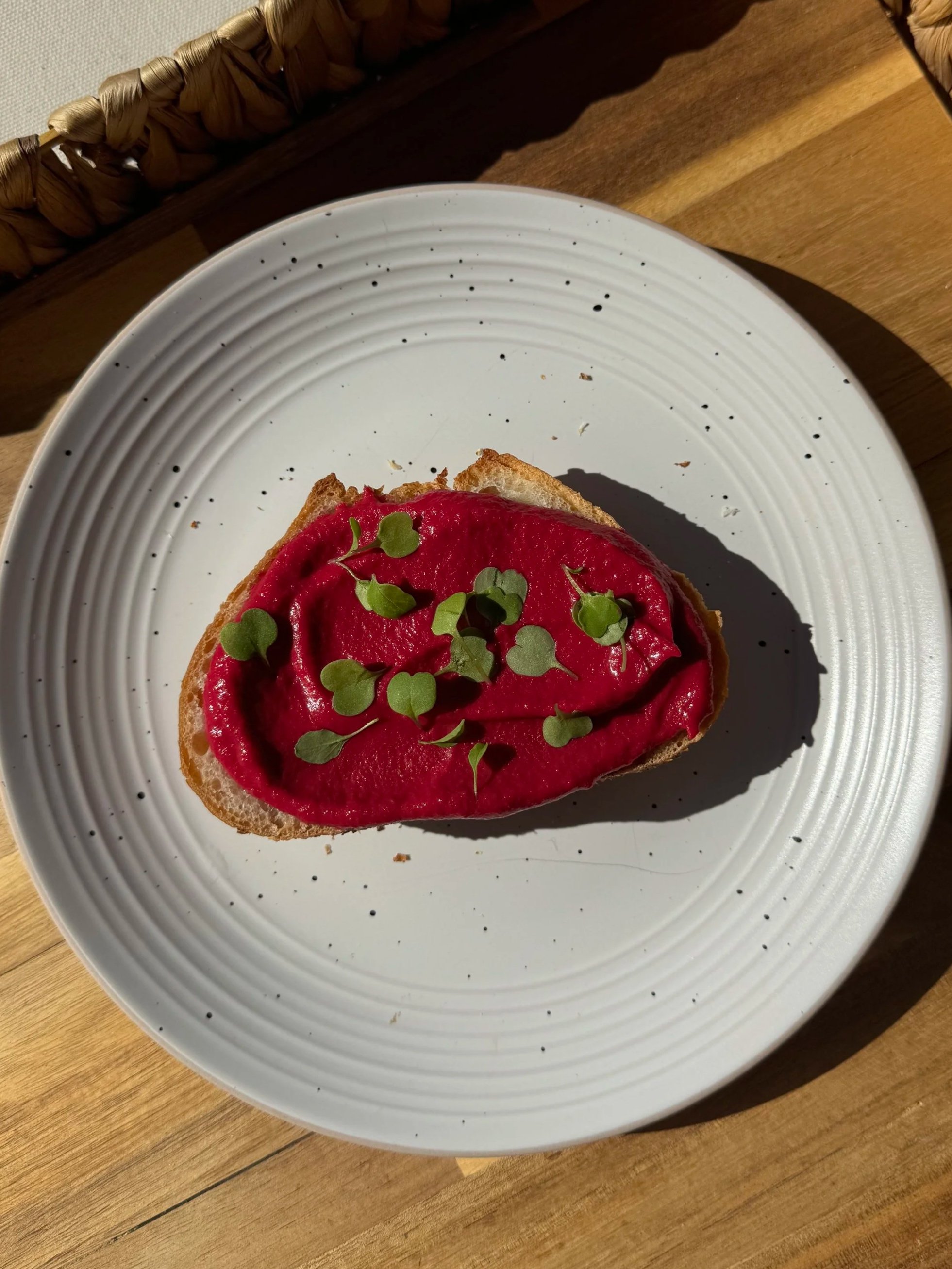 A slice of bread topped with a beet hummus and garnished with small fresh green herbs on a white plate with a textured rim, placed on a wooden surface.