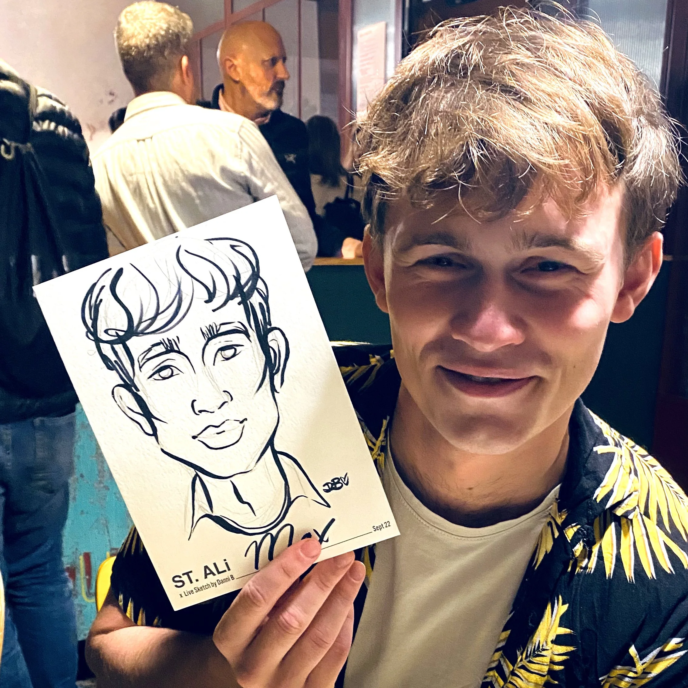 A person smiling while holding a caricature drawing of themselves, with people in the background at a social event.
