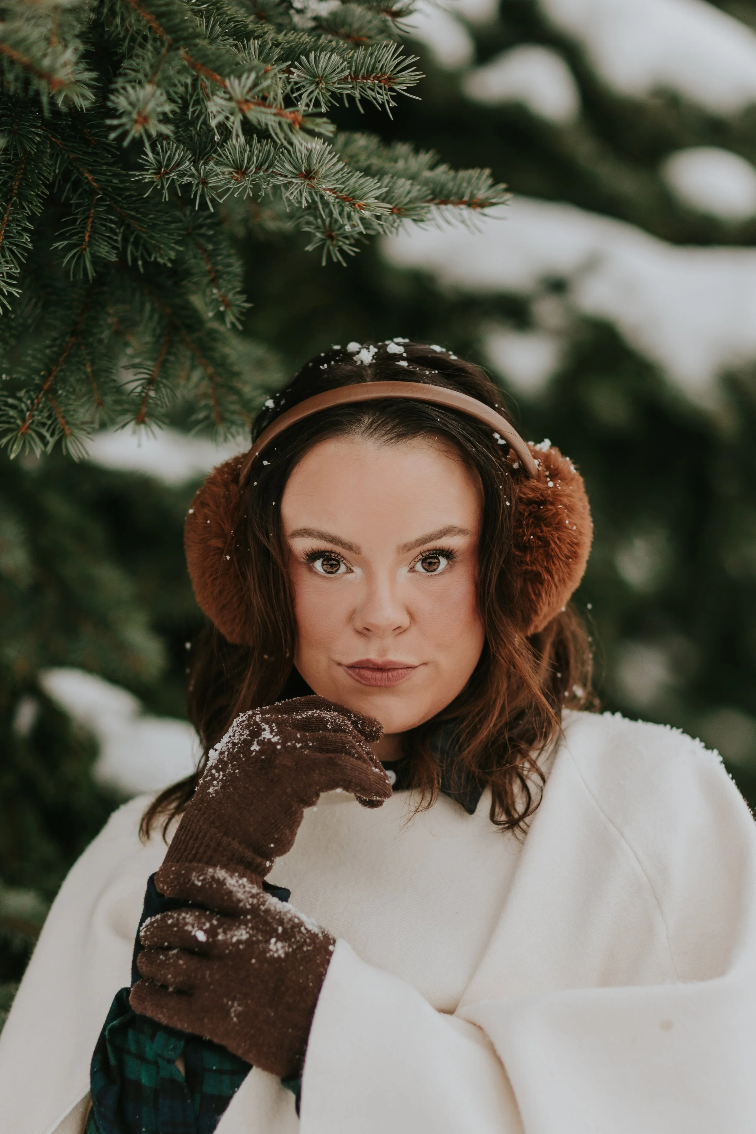 A woman wearing brown earmuffs and gloves, a white coat, and a black shirt with a collar, standing outdoors in snow-covered trees with green pine branches.