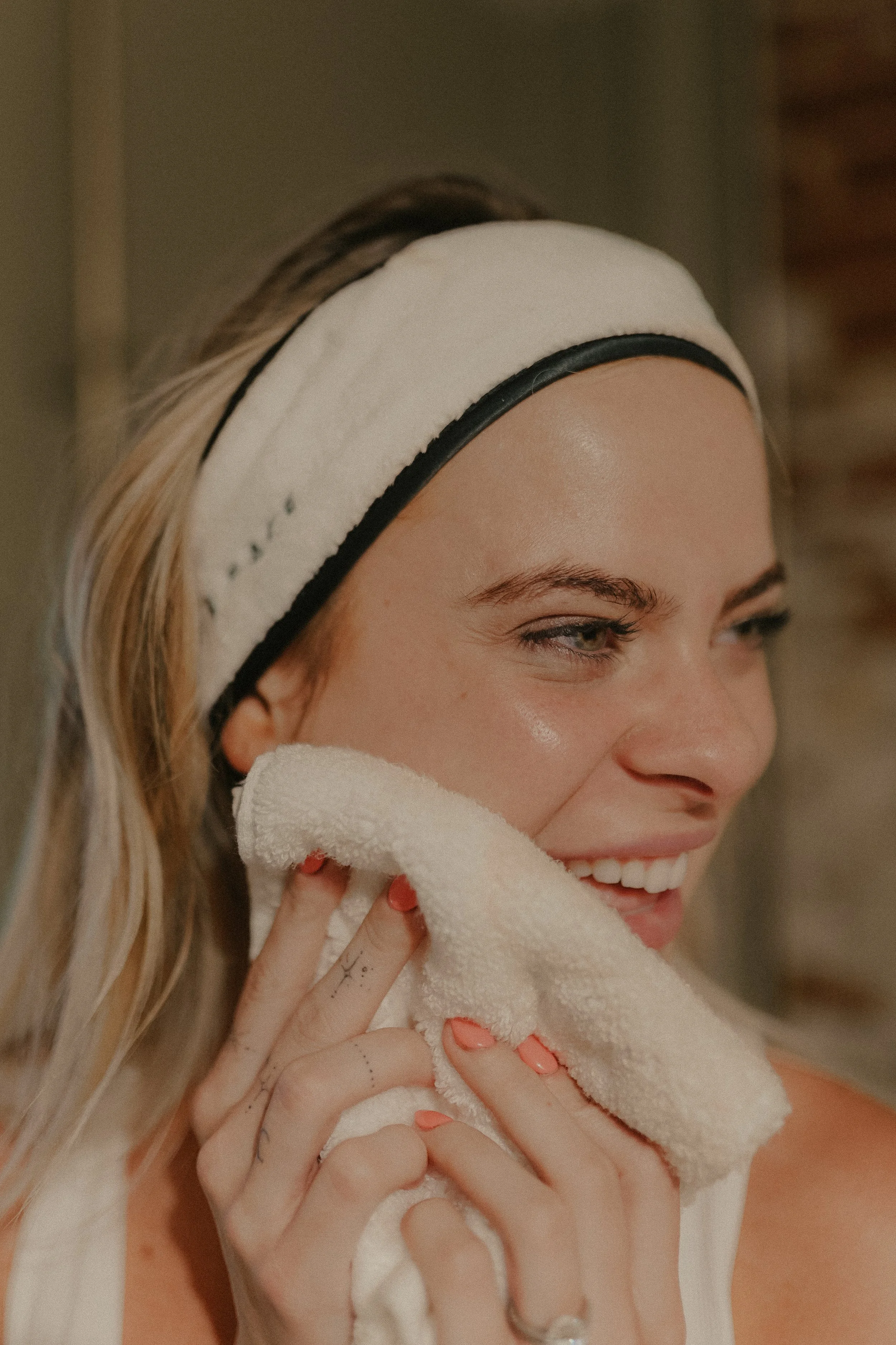 A woman with blonde hair wearing a white headband, smiling and touching her face with a towel.