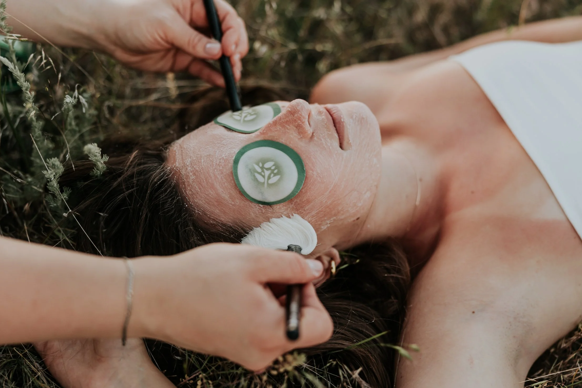 A woman receiving a facial treatment with a green facial mask, smiling with eyes closed, while aesthetician applies mask with brushes.