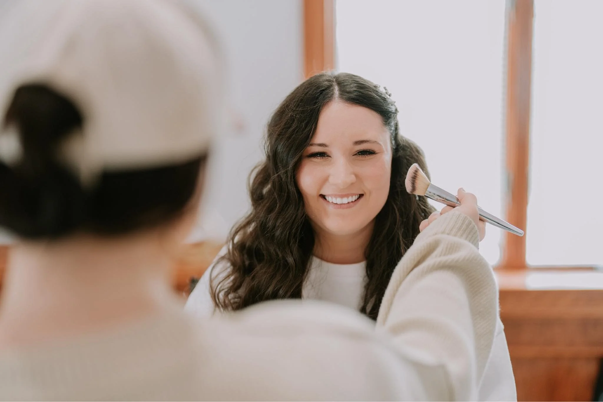 A makeup artist applying eyeshadow to a woman with closed eyes, sitting in a chair, while the artist laughing and holding an eyeshadow palette.