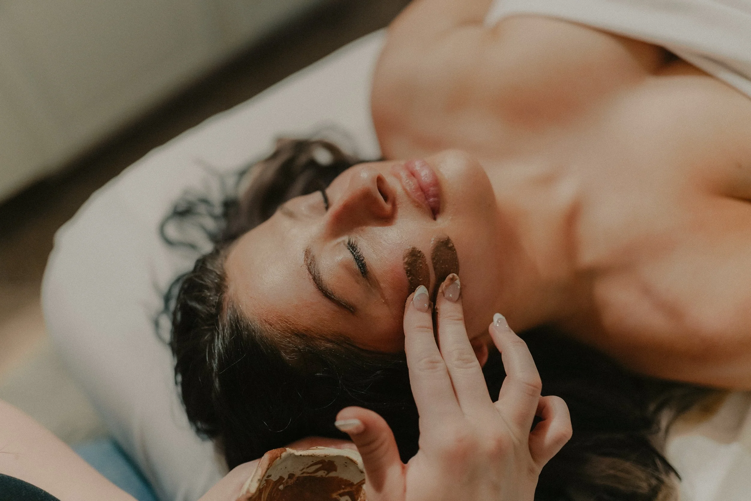 A woman lying on a bed with her eyes closed, receiving a facial treatment with a mud mask applied to her eyelids.