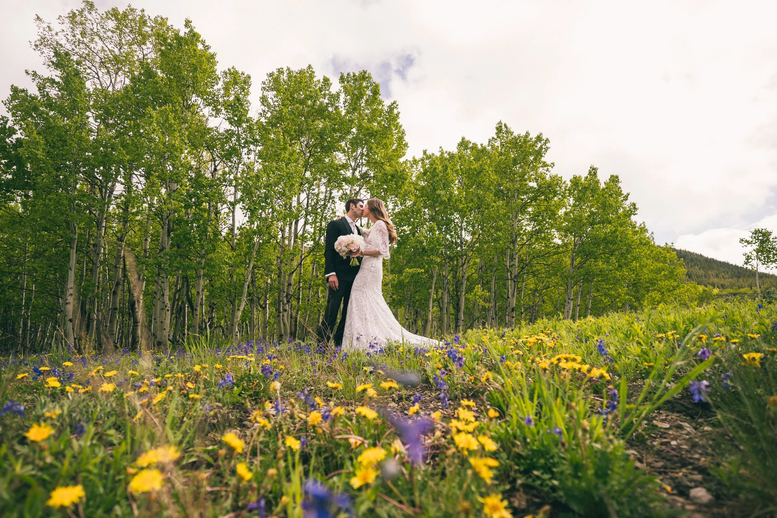 Bride and groom kissing in a field of wildflowers with a backdrop of green trees.