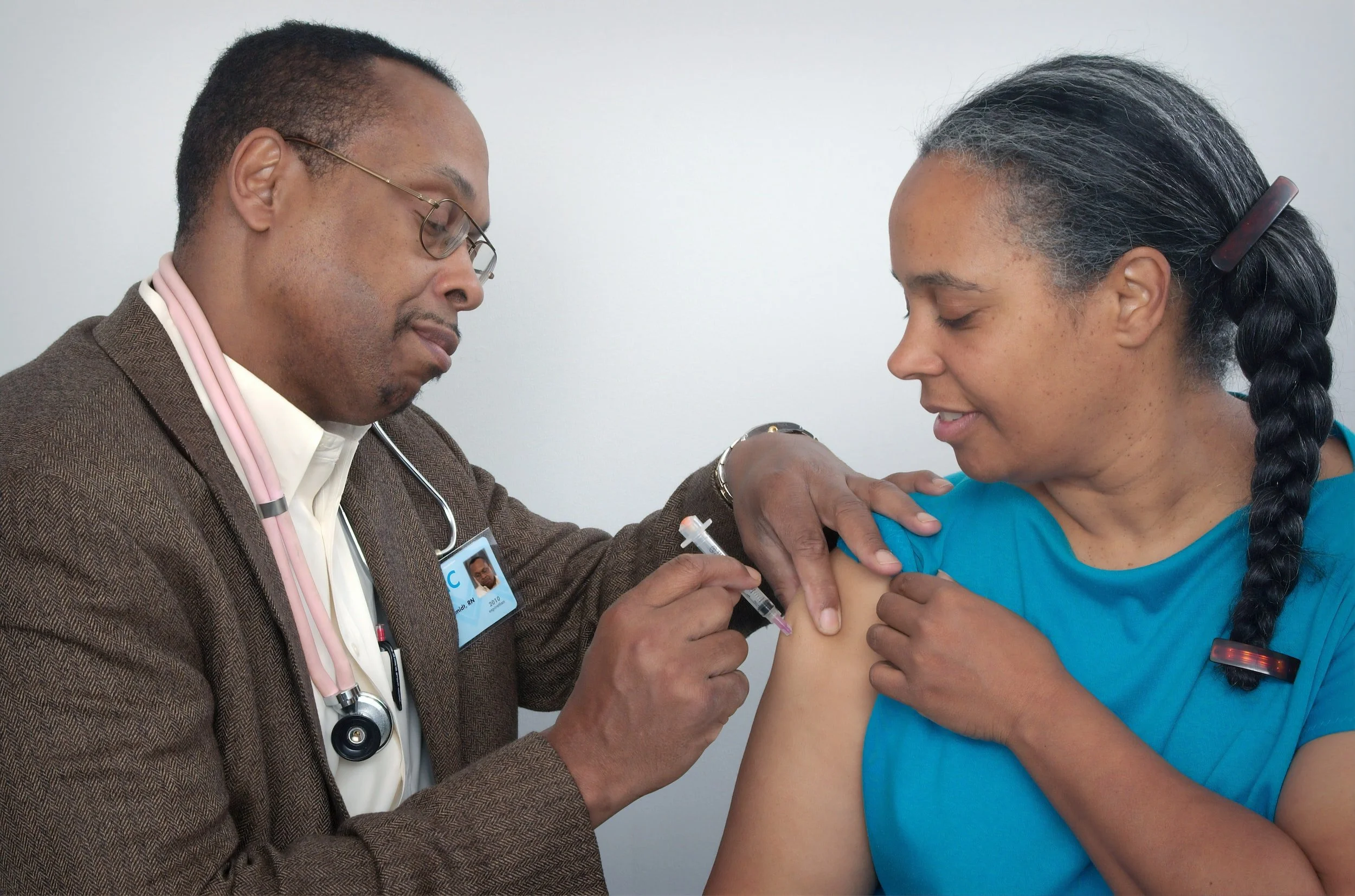 A healthcare professional is administering a vaccine to a woman in a blue shirt. The woman is smiling and the healthcare professional is wearing a stethoscope.