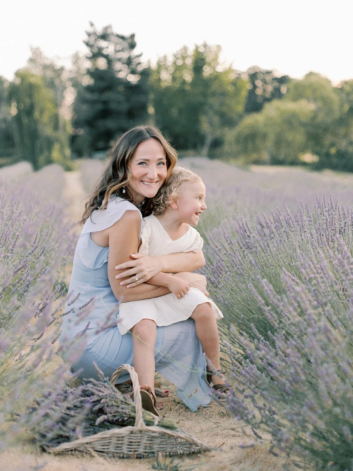 A woman and a young girl sitting together in a lavender field, smiling and enjoying the moment. The woman has wavy brown hair and is wearing a light blue dress, while the girl has curly blonde hair and is wearing a cream-colored dress. The background features tall trees and a clear sky.