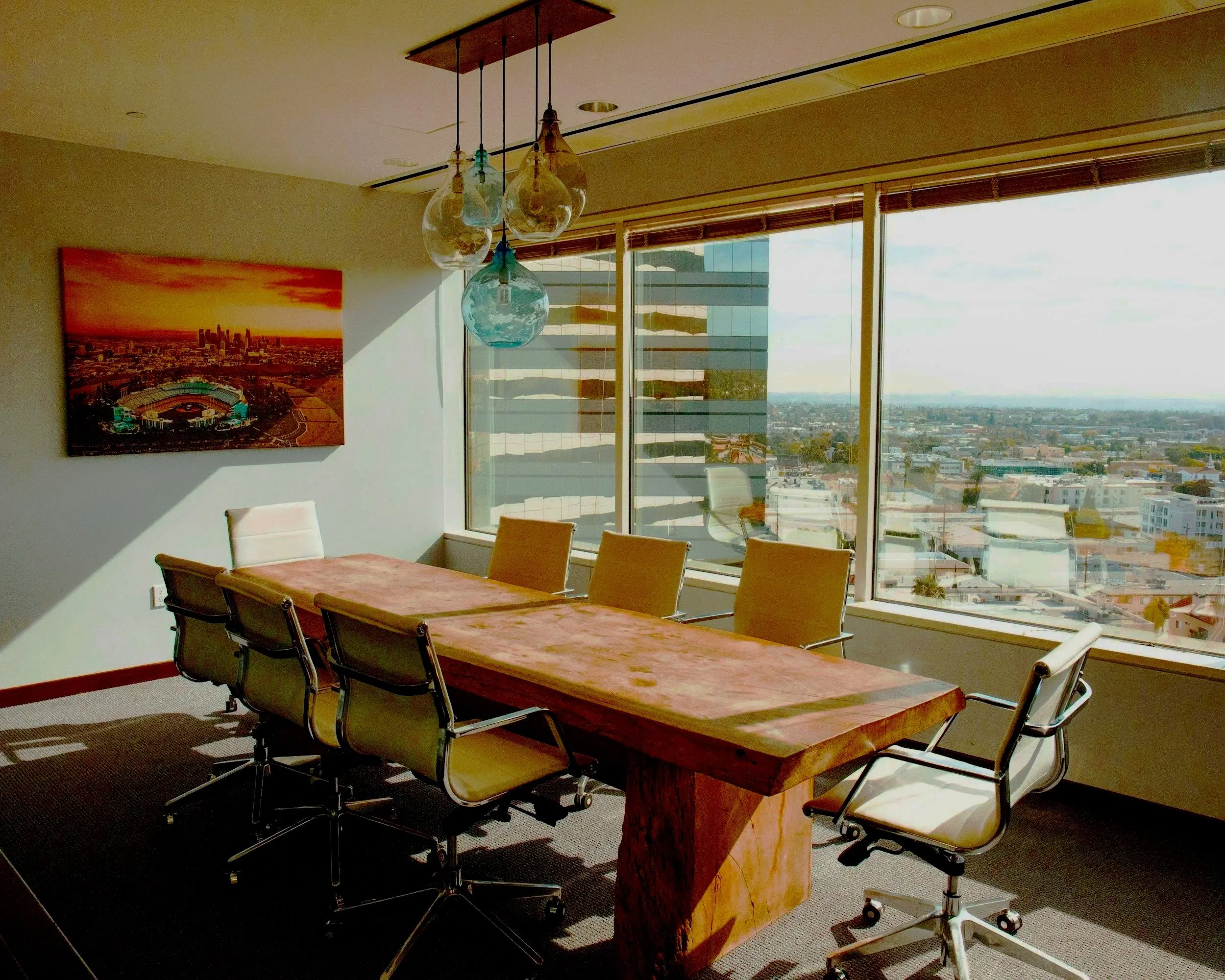 Modern conference room with a large wooden table, six yellow office chairs, a colorful abstract artwork on the wall, hanging glass pendant lights, and large windows with city views.