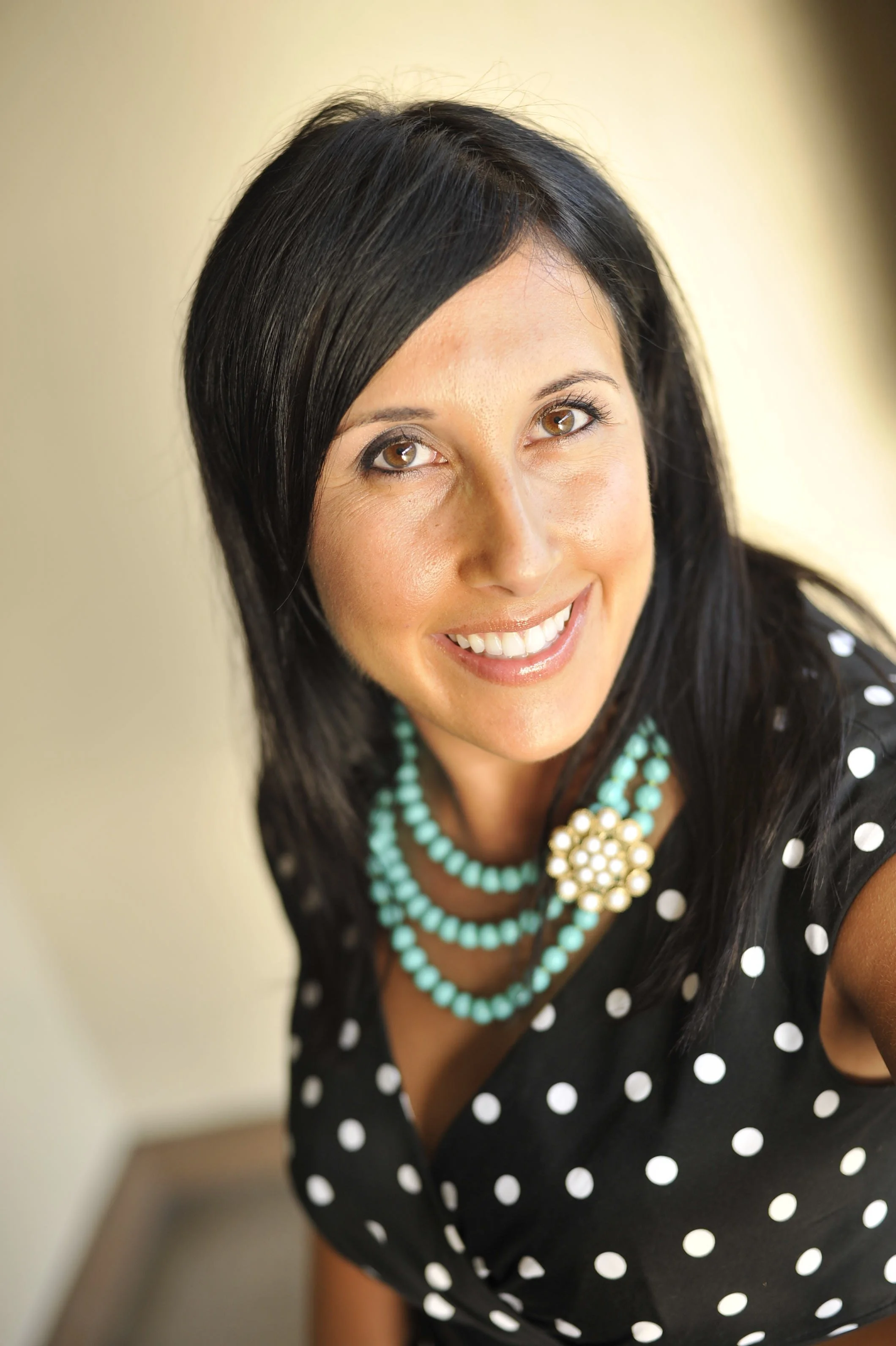 A woman with dark hair wearing a black dress with white polka dots, turquoise jewelry including a multi-strand necklace and a brooch, smiling at the camera.