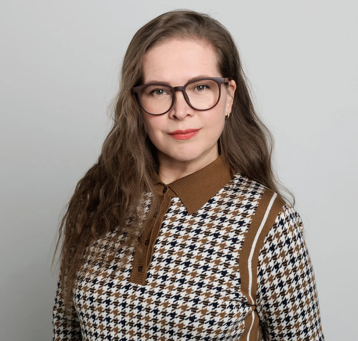 A woman with long, wavy brown hair and glasses, wearing a brown and white houndstooth patterned shirt with a brown collar, posing against a plain light gray background.