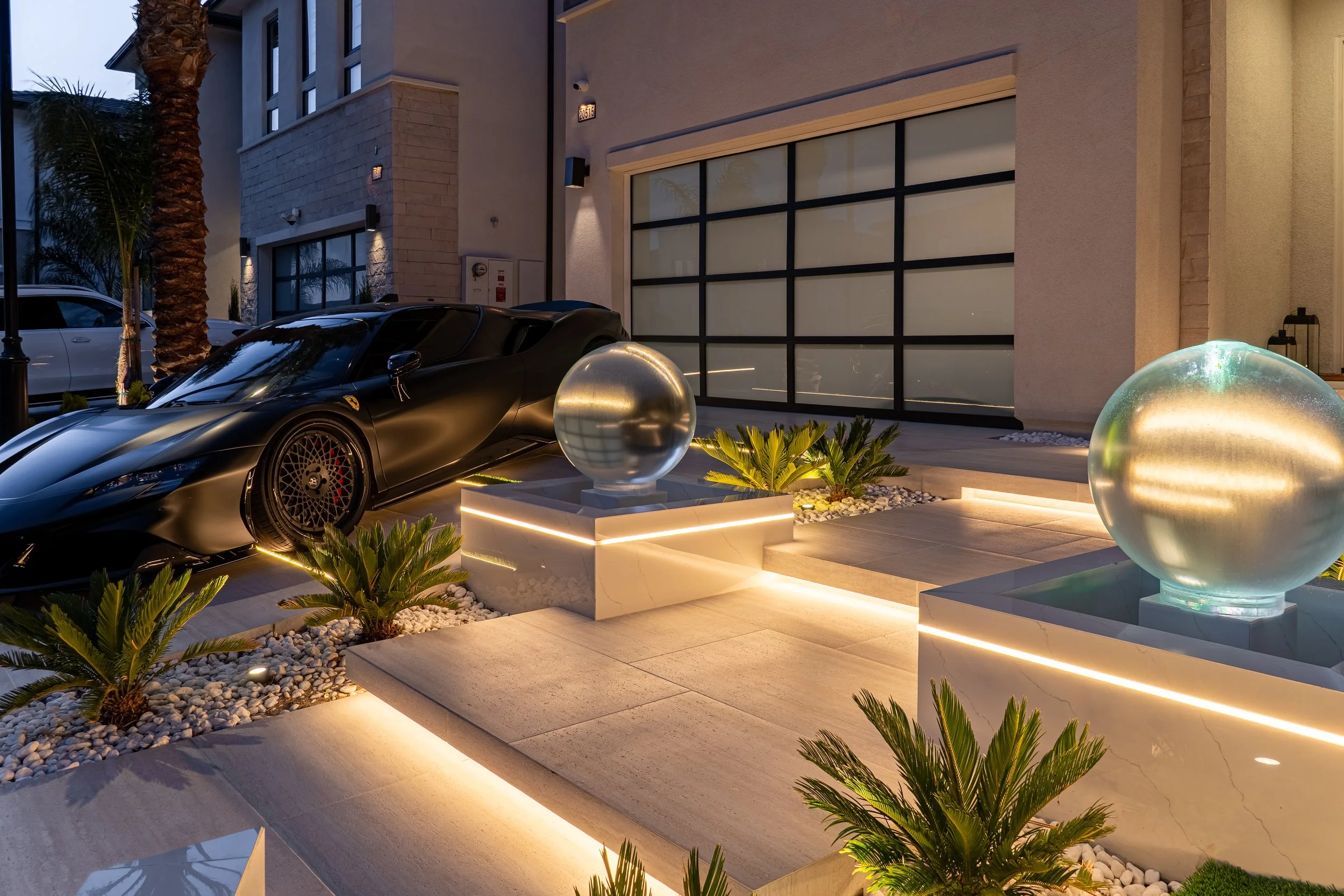 Luxury modern house with a black sports car parked in front, illuminated sculptures, palm trees, and LED lighting accents.
