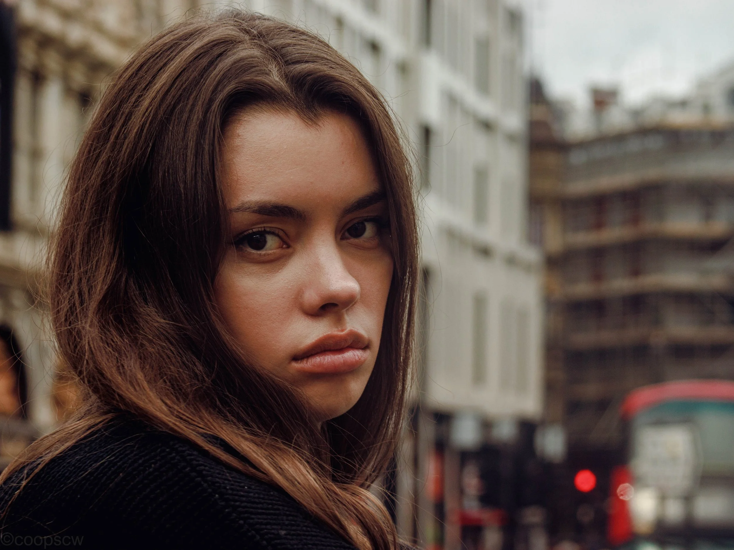 Street portrait of a young woman looking towards the camera, with London buildings and a red bus behind her.