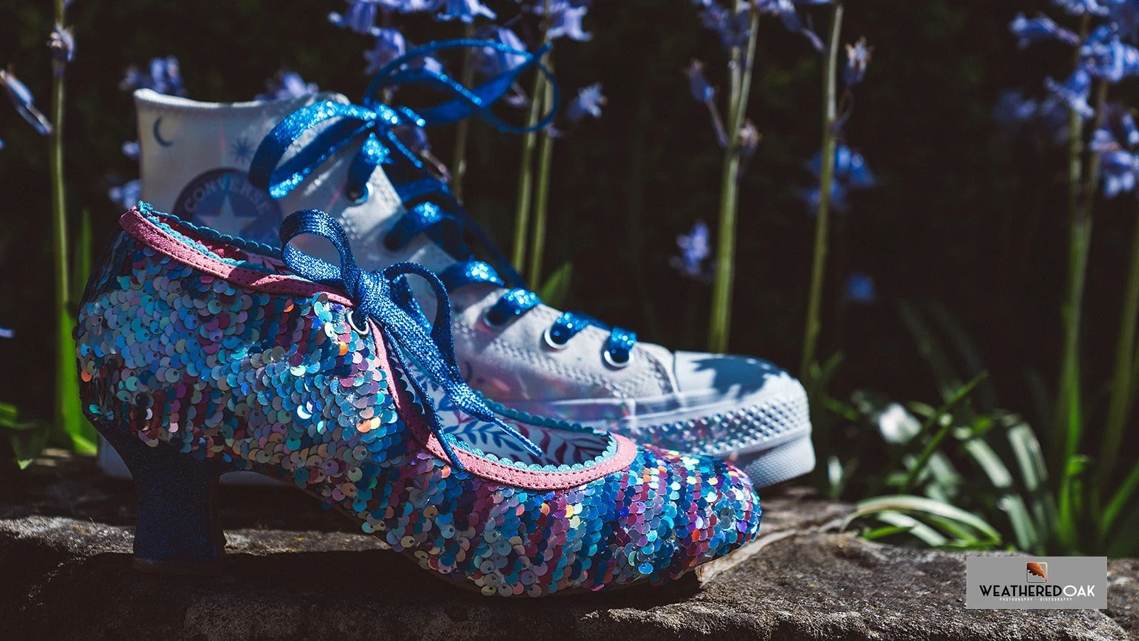 A high-heeled shoe covered in blue sequins with blue laces is placed next to a white high-top sneaker with blue laces, in a garden setting with purple flowers in the background.