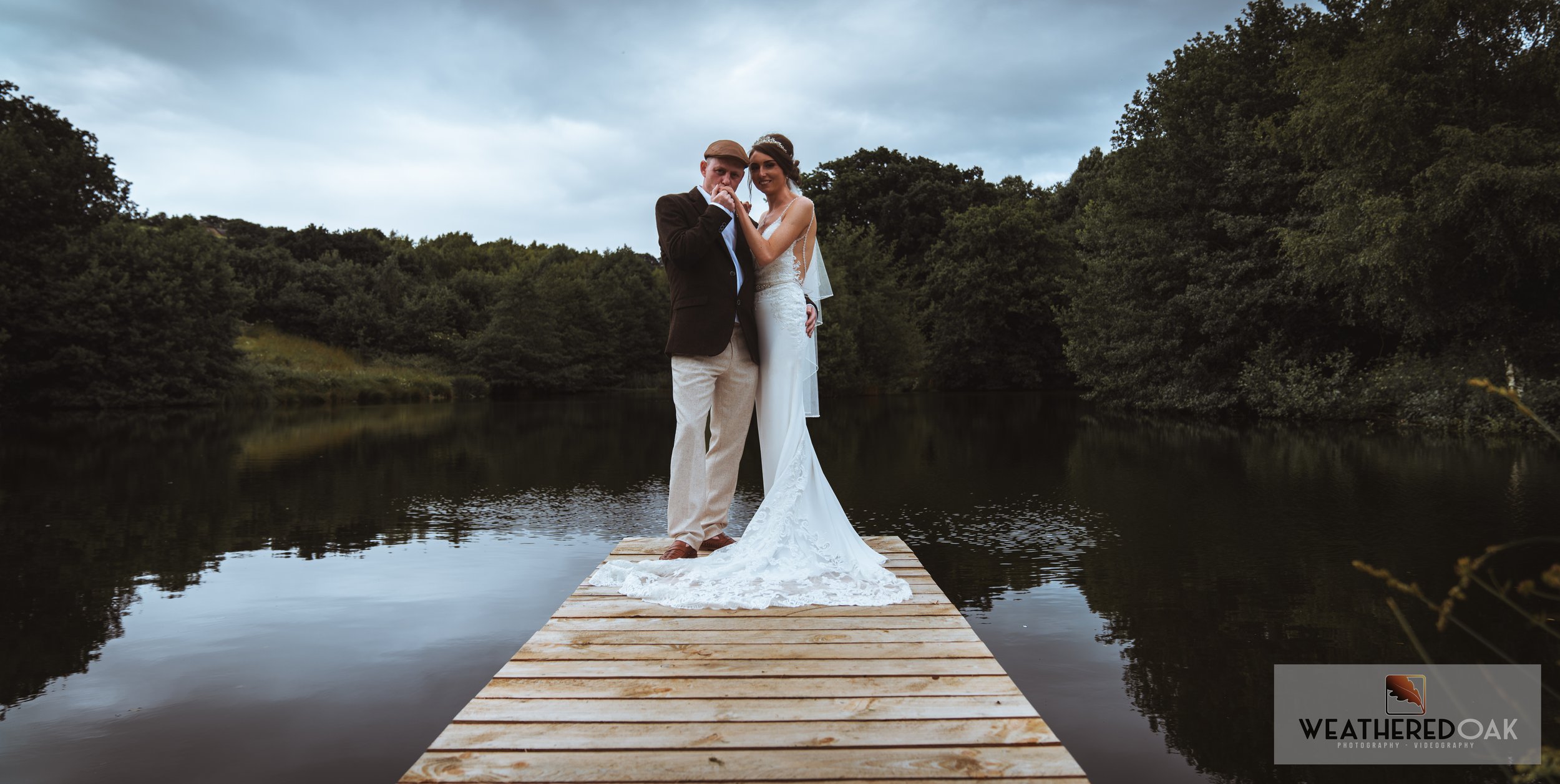 Bride and groom standing on a wooden dock over a lake surrounded by dense greenery, cloudy sky overhead.