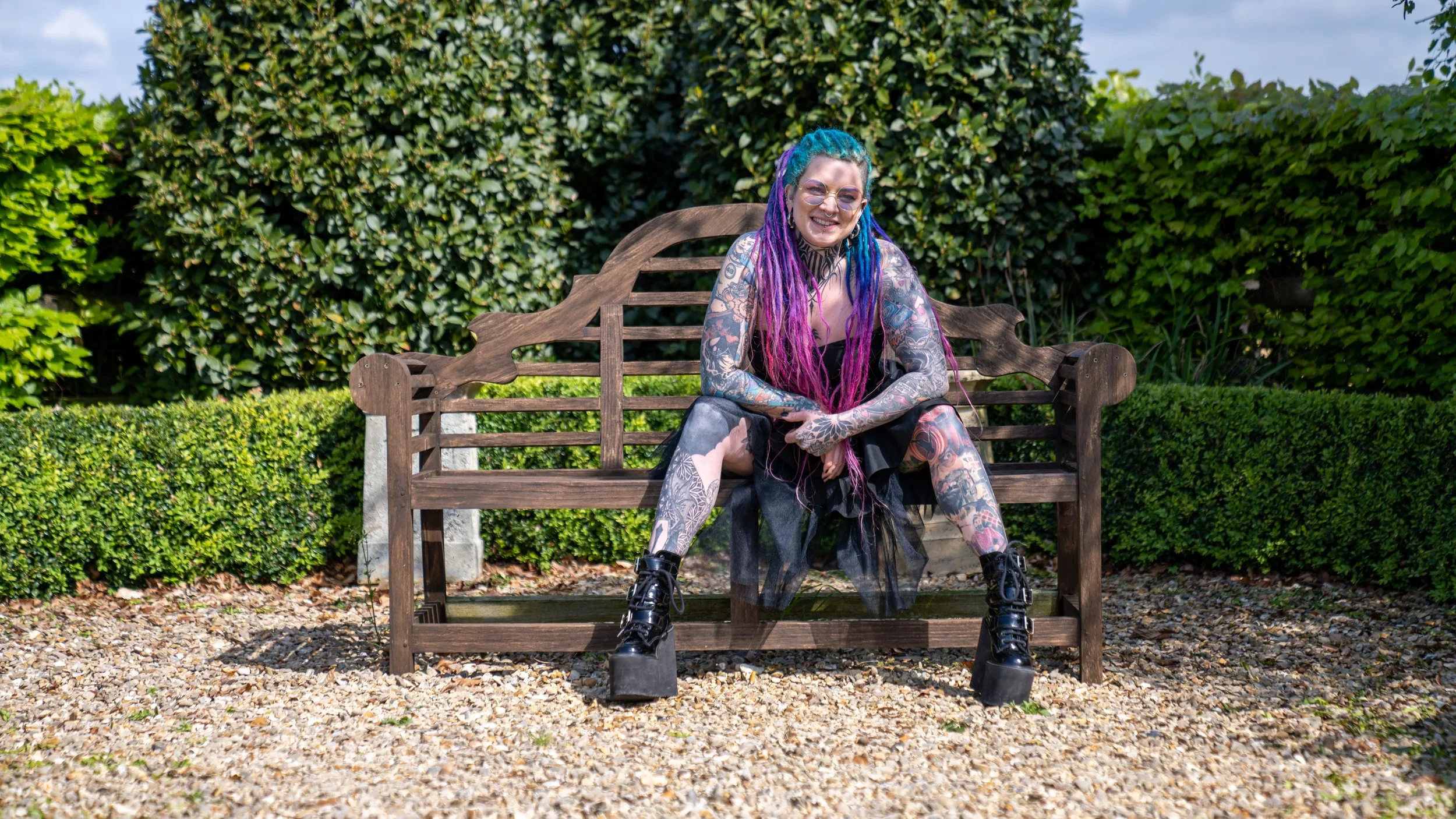 Person with colorful dreadlocks and tattoos sitting on a wooden bench in a garden.