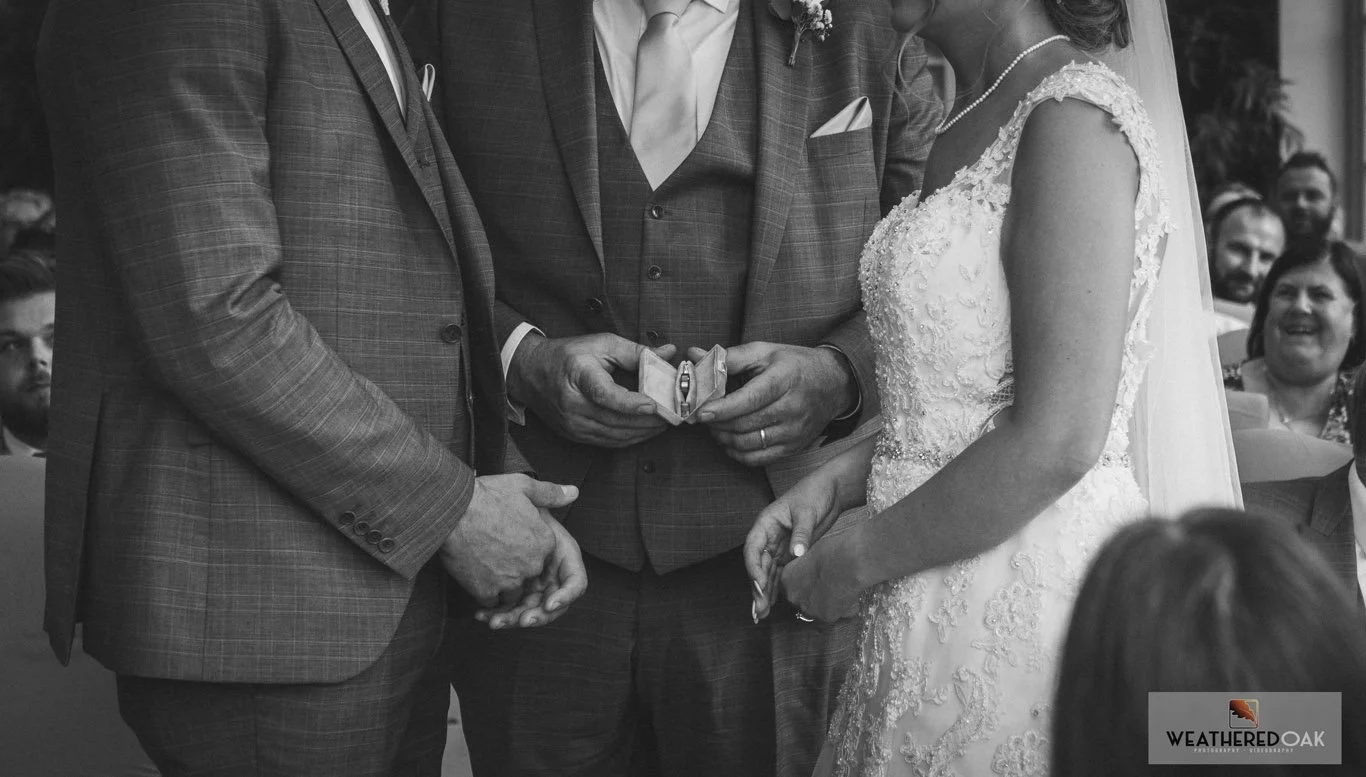 A black and white image of a couple dressed in wedding attire during a wedding ceremony, featuring a person holding a ring box.