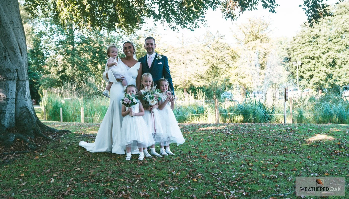 A wedding photo with a bride and groom standing on a lawn under a tree. The bride holds a baby while three young flower girls in white dresses stand in front, holding flower bouquets. Trees and greenery are visible in the background.