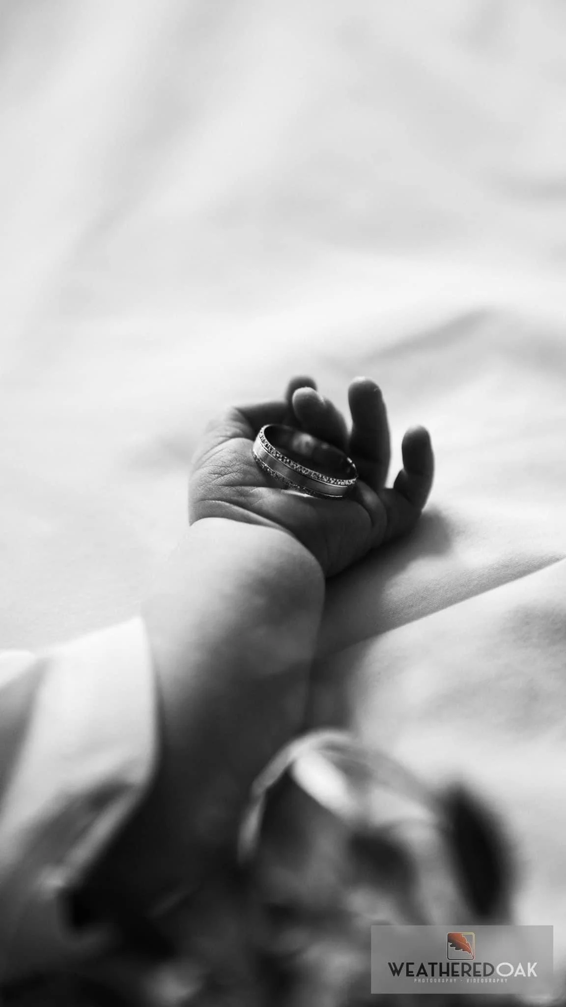 Black and white photo of a baby's hand holding a ring on a fabric surface.