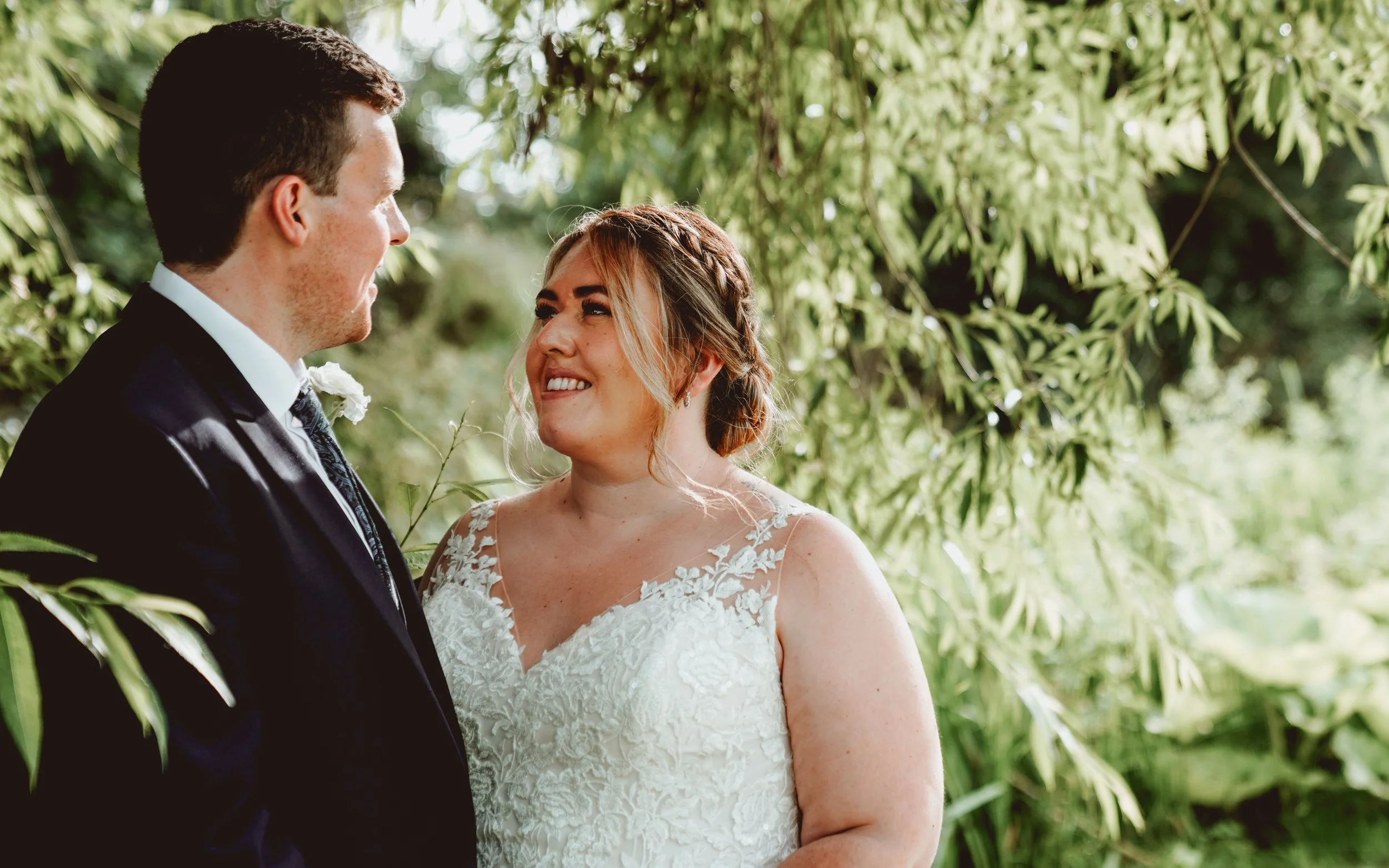 Bride and groom smiling at each other under leafy trees
