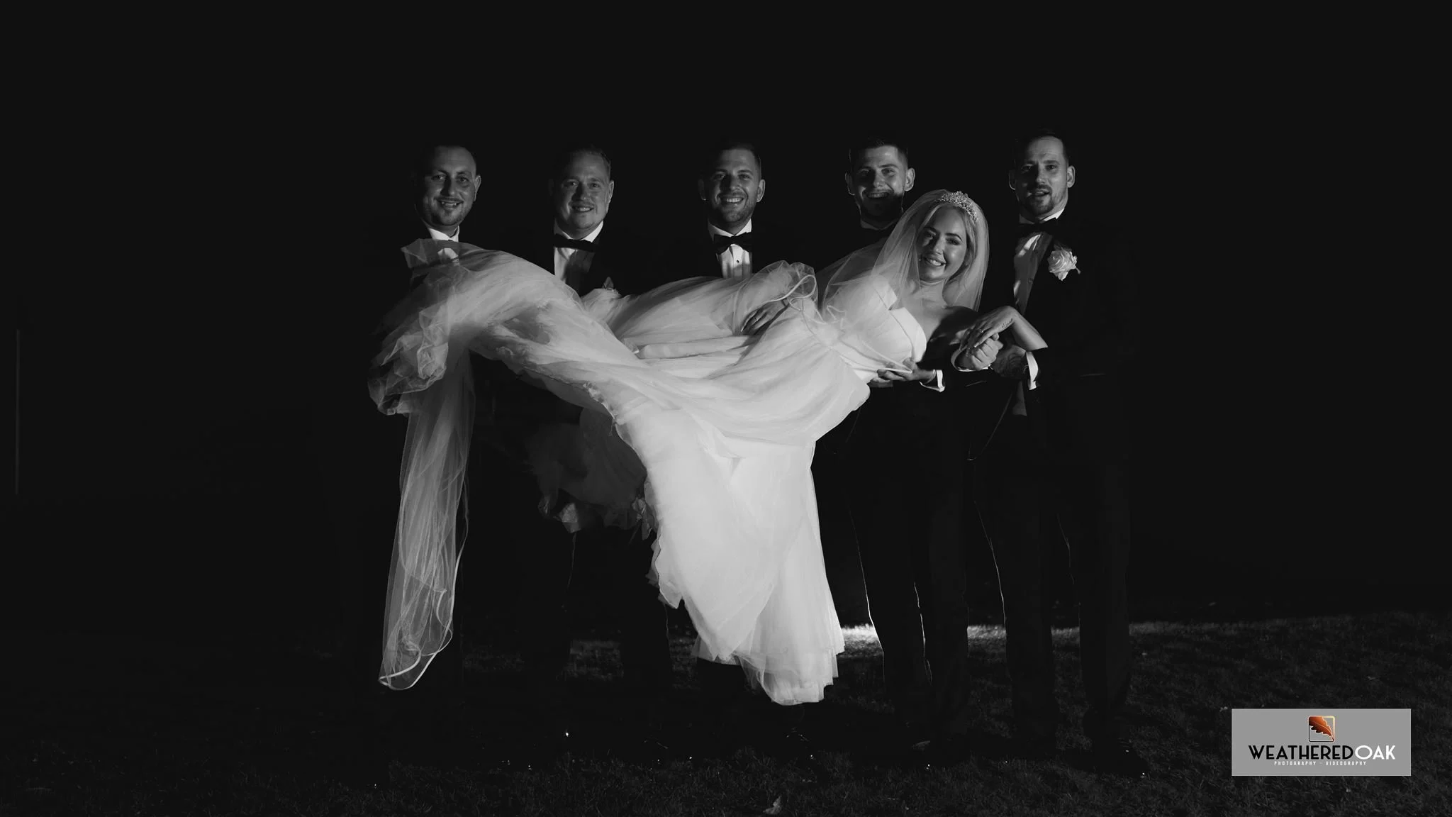 Black and white photo of five men in suits and a bride in a wedding dress. The men are holding the bride horizontally, posing and smiling.