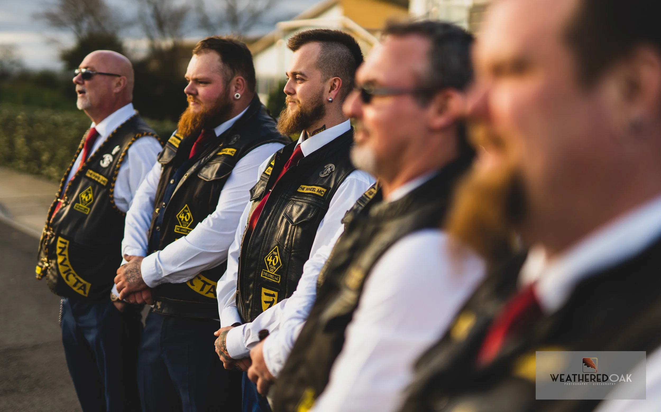 Group of men in black leather vests standing outdoors, wearing white shirts and red ties, with patches on vests; logo of "Weathered Oak" in corner.