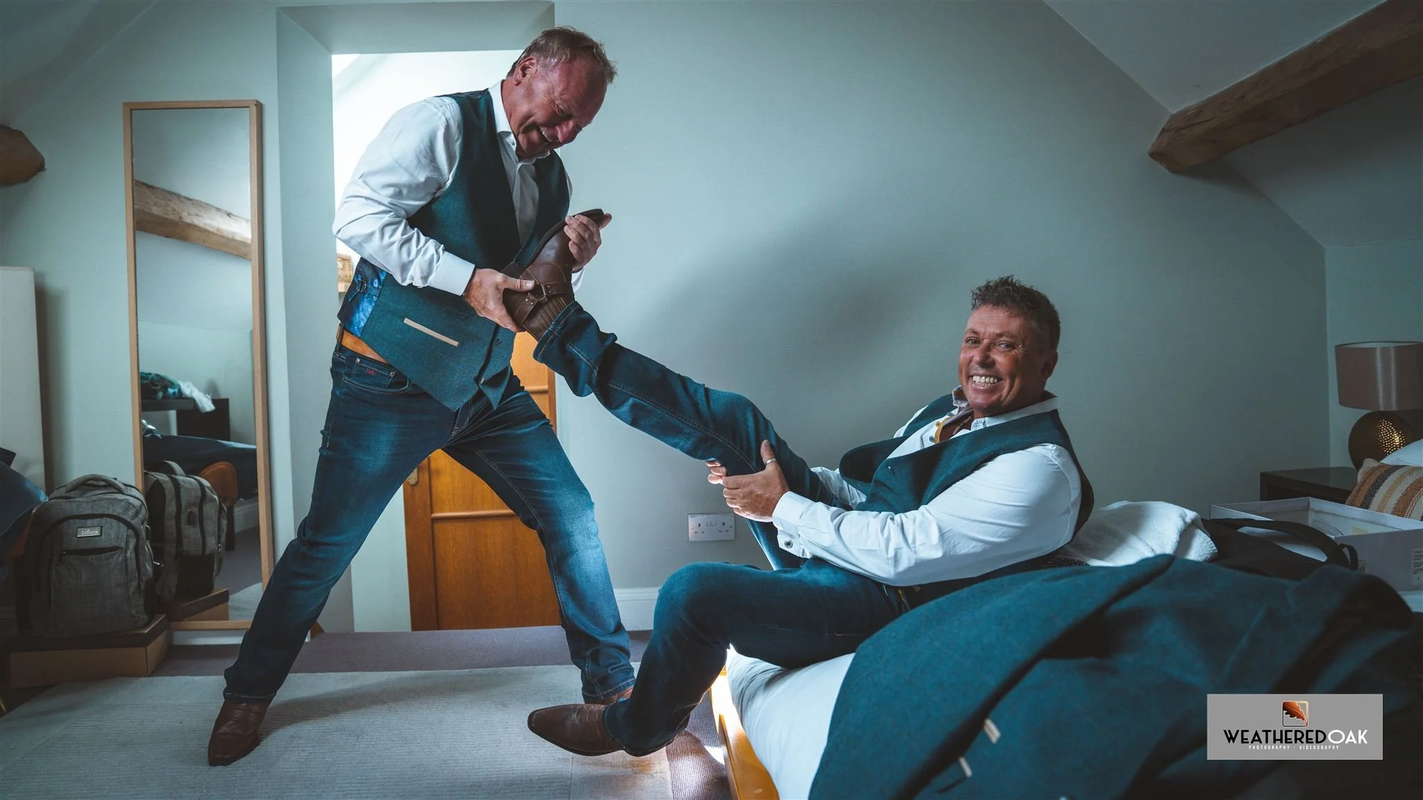 Two men in matching vests and jeans, one sitting on a bed holding his leg, while the other pulls on his boot with a playful, laughing expression.