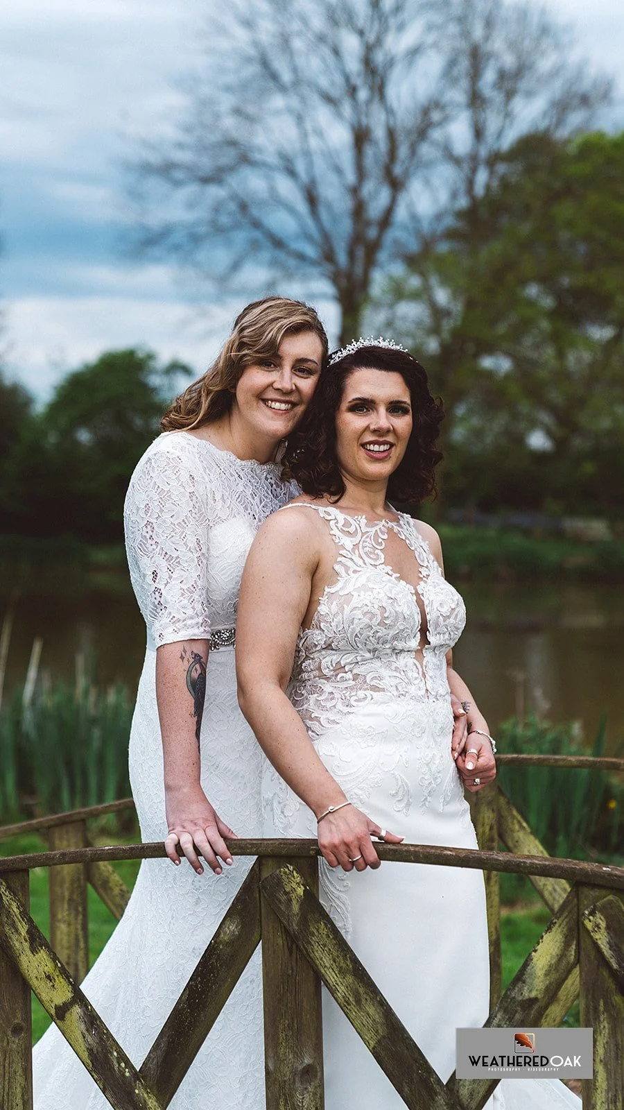 Two brides in white wedding dresses standing outdoors on a wooden bridge, smiling, with trees and a lake in the background.