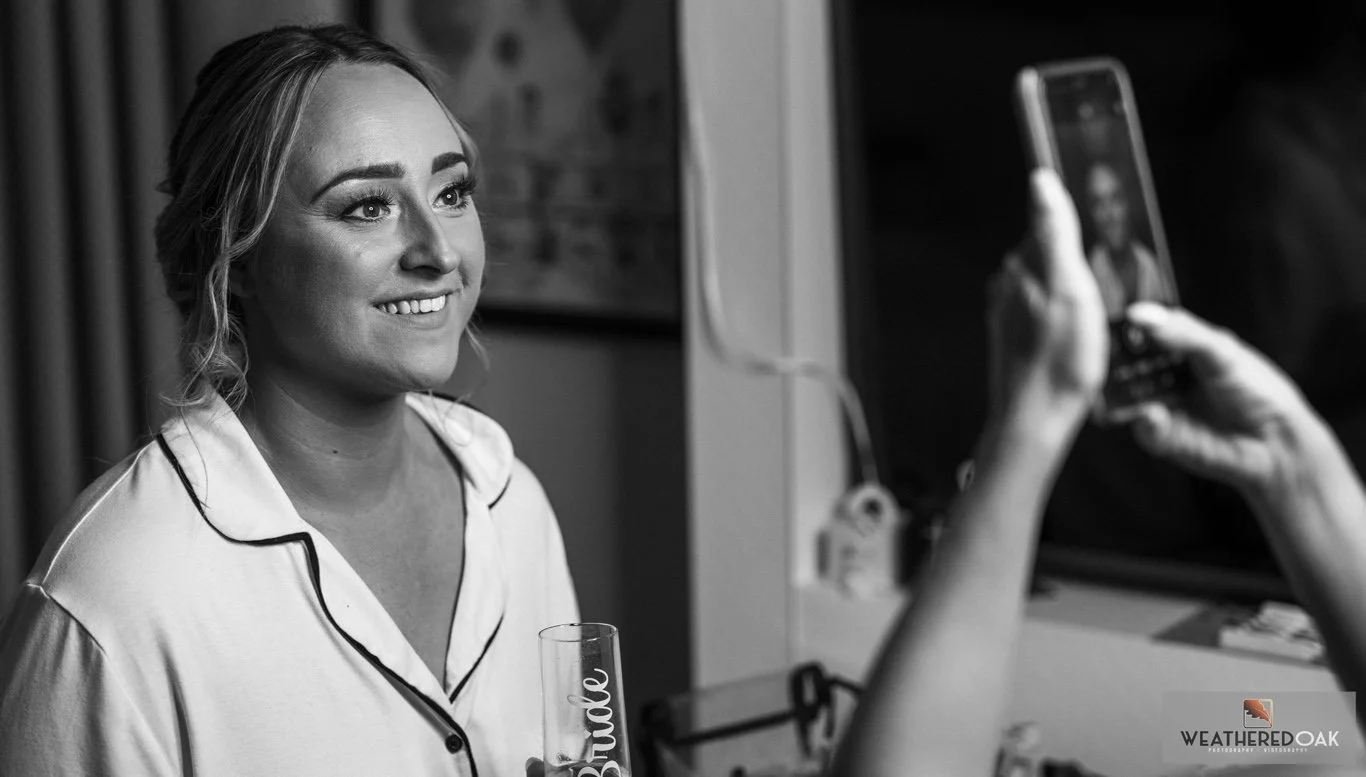 Black and white photo of a person smiling at a camera, holding a glass with 'bride' written on it. Another person is taking a photo of them with a smartphone.