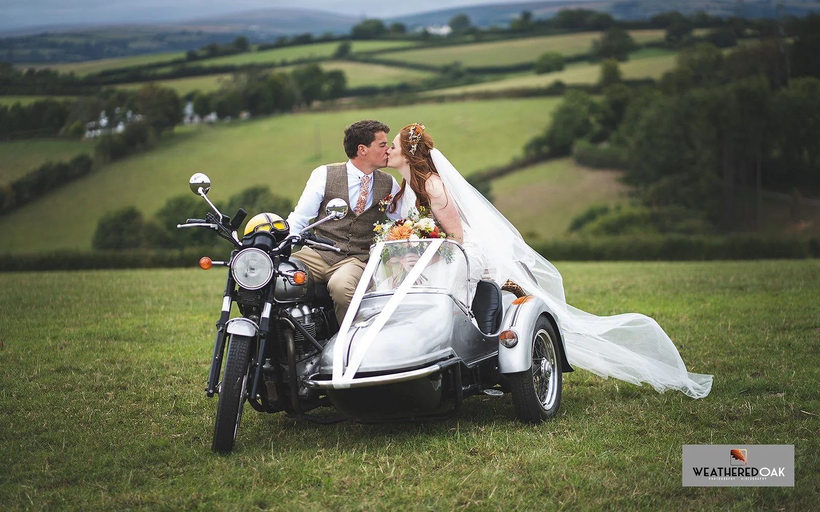 Bride and groom kissing while sitting on a motorcycle with a sidecar in a scenic countryside setting.