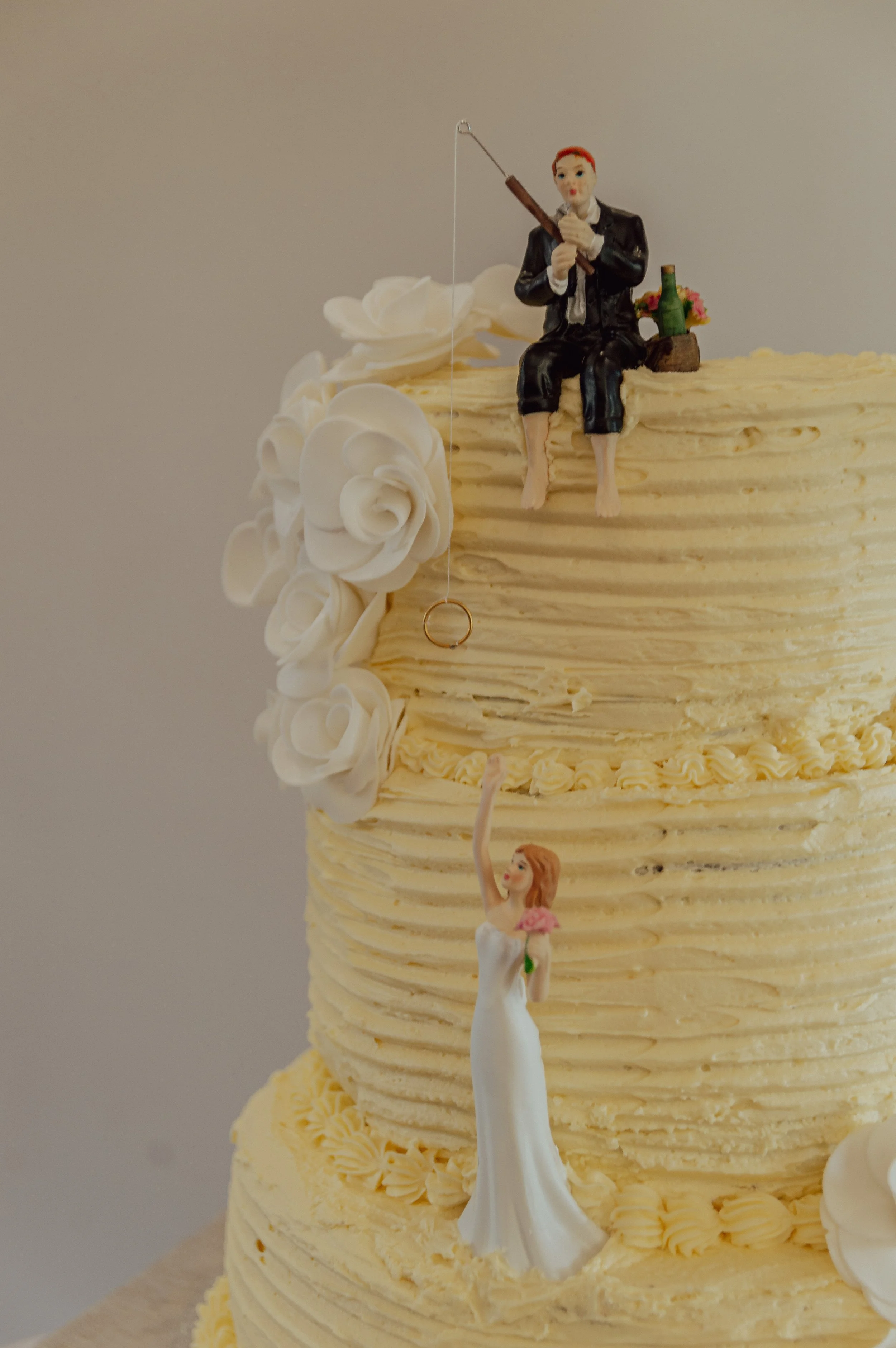 Wedding cake with white frosting, featuring a figurine of a groom fishing and a bride reaching up, adorned with edible roses.