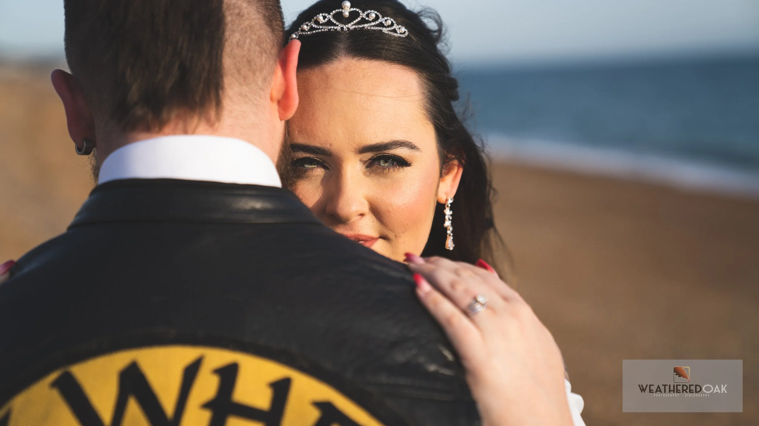 Woman with tiara embracing person wearing a leather jacket on a beach.