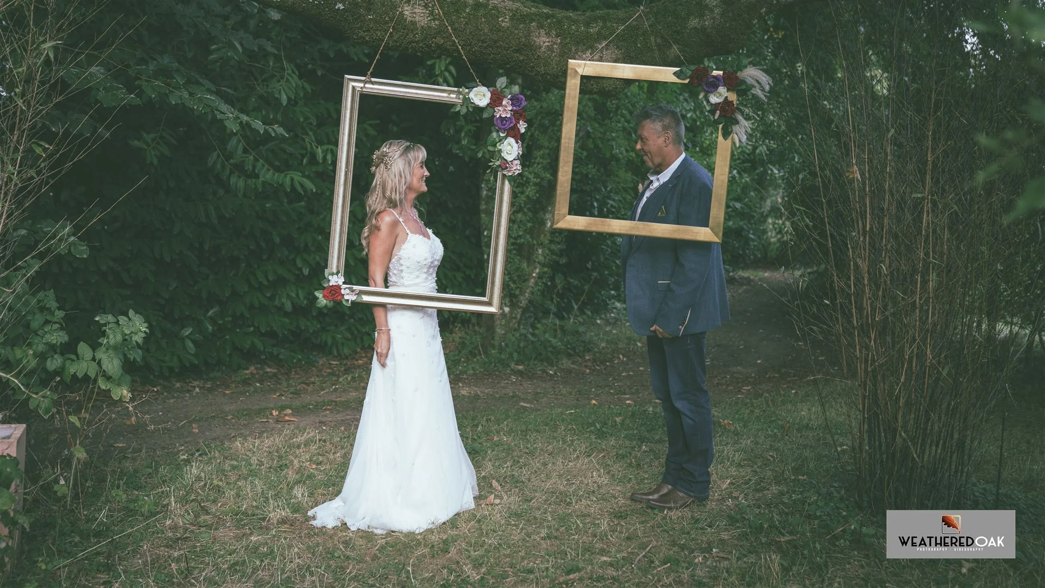 Bride and groom standing in outdoor setting, each framed by a hanging picture frame adorned with flowers, looking at each other. The frames are gold and silver.
