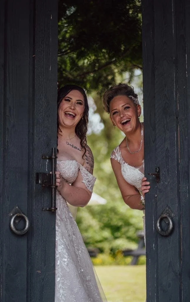 Two women in wedding dresses smiling and peeking through open wooden doors with greenery in the background.