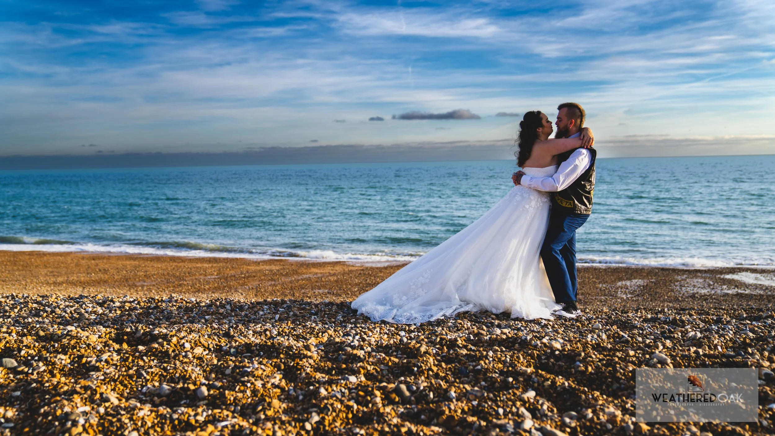 Bride and groom embracing on a pebble beach with ocean backdrop under blue sky.