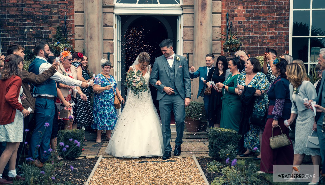 A wedding couple exits a building surrounded by guests throwing confetti, with the bride in a white gown holding flowers and the groom in a gray suit. Guests line the pathway, celebrating the newlyweds.
