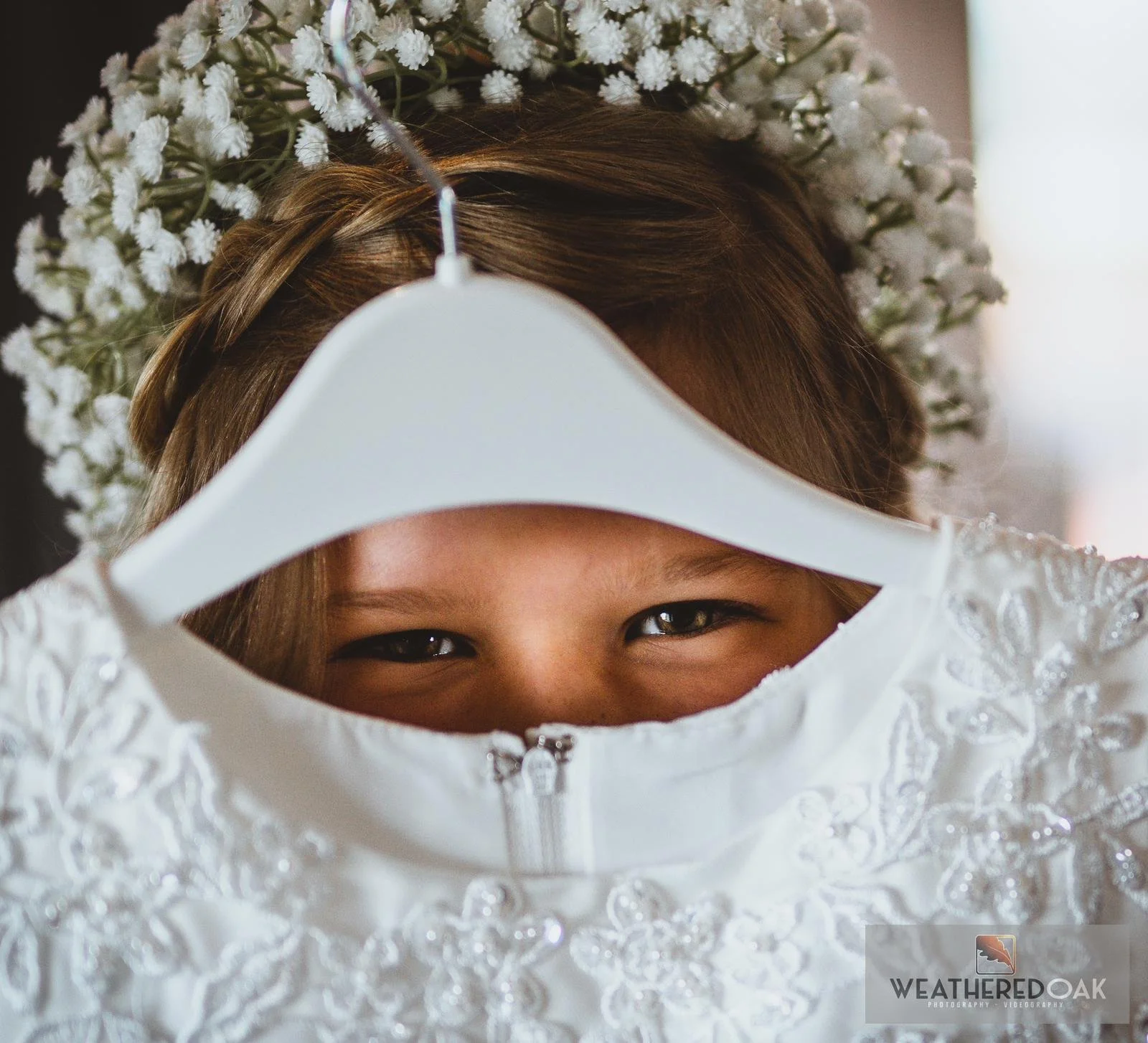 Smiling child holding white dress on a hanger, with flower crown made of baby's breath.