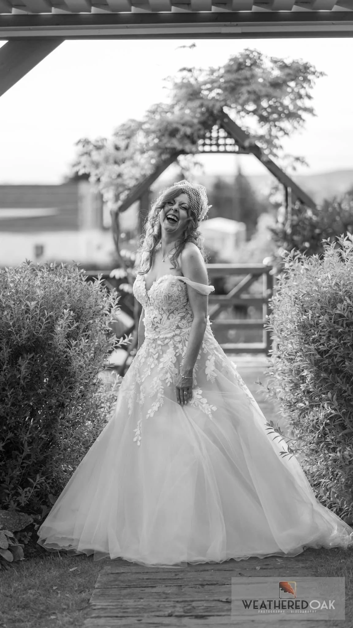 Smiling bride in white lace wedding dress in garden setting, black and white photo.