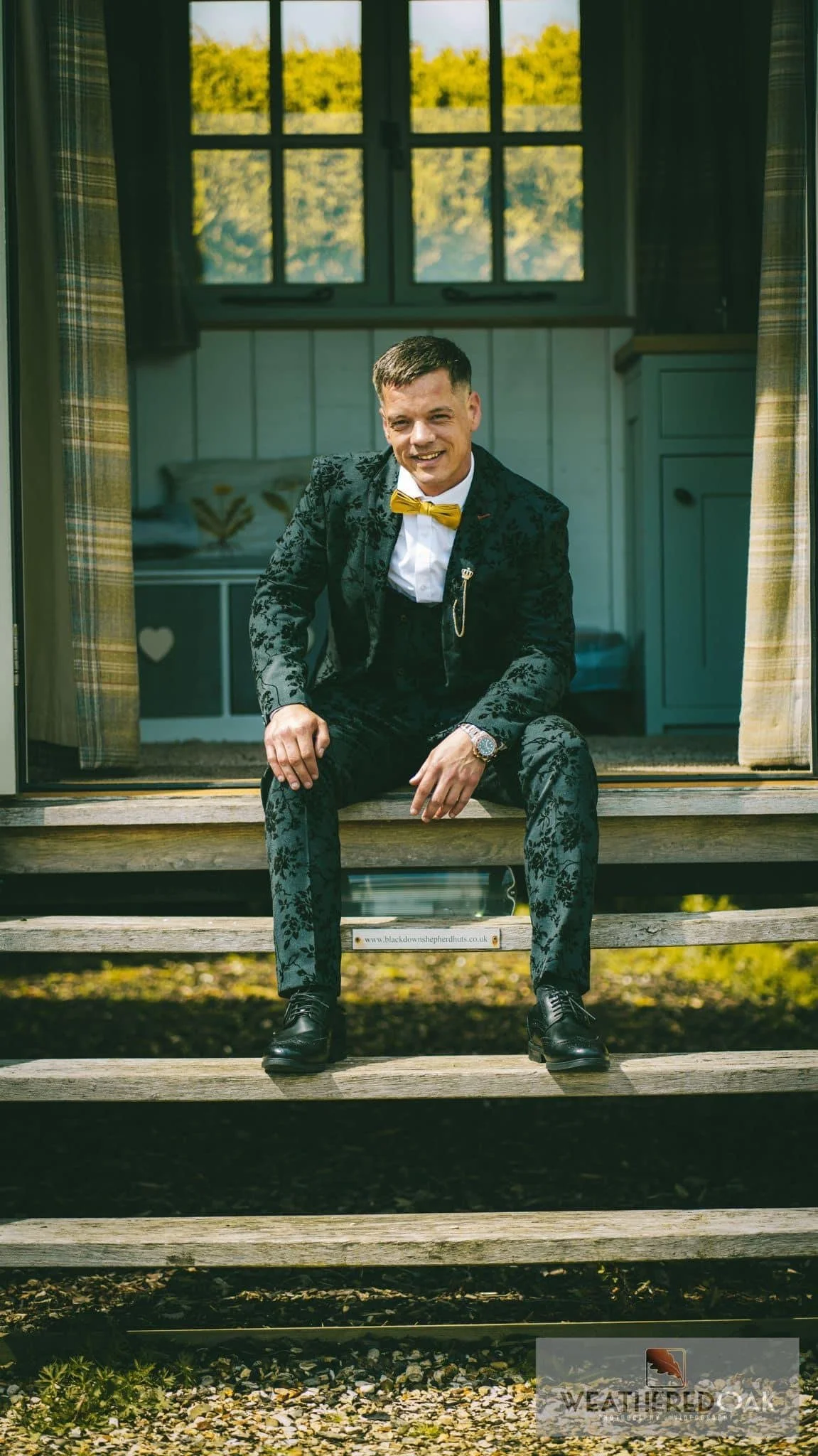 Man in floral suit sitting on wooden steps in front of a cabin.