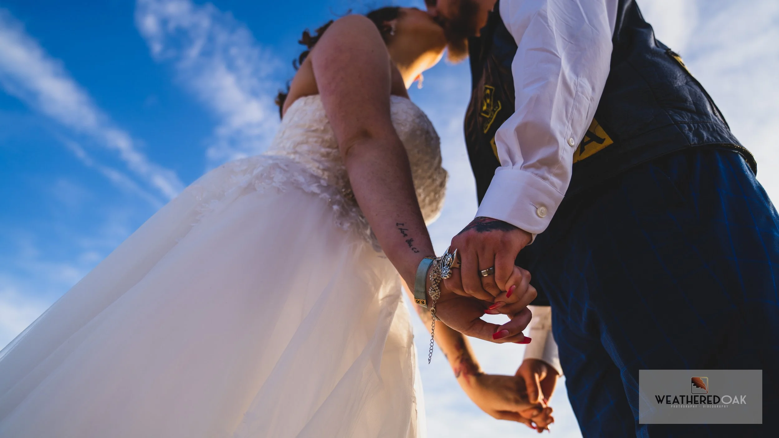Low angle view of a couple holding hands and kissing, with the bride in a white dress and the groom in a suit and vest, set against a blue sky background.