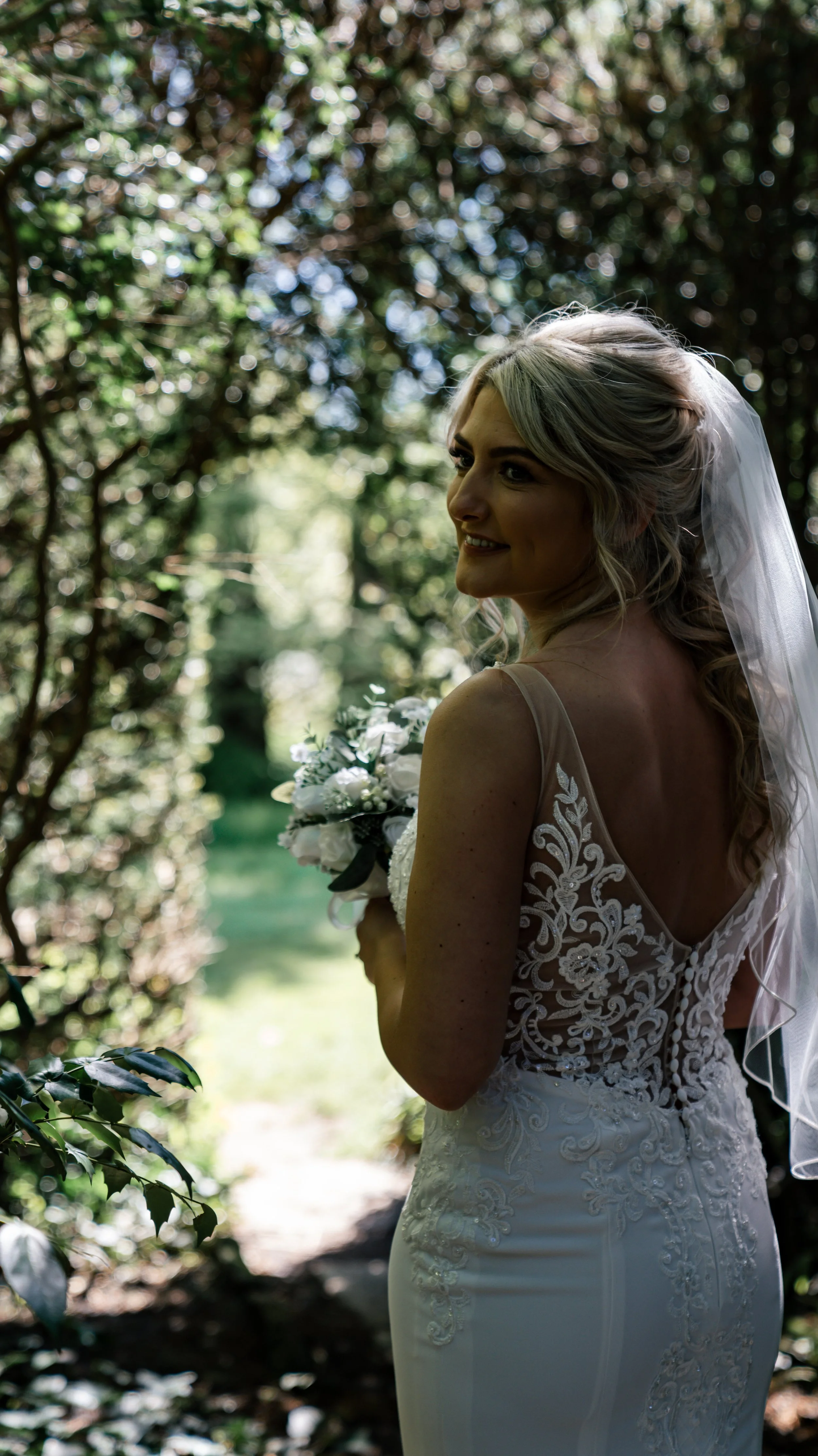 Bride in lace wedding dress with veil, holding bouquet, standing in sunlight surrounded by greenery.