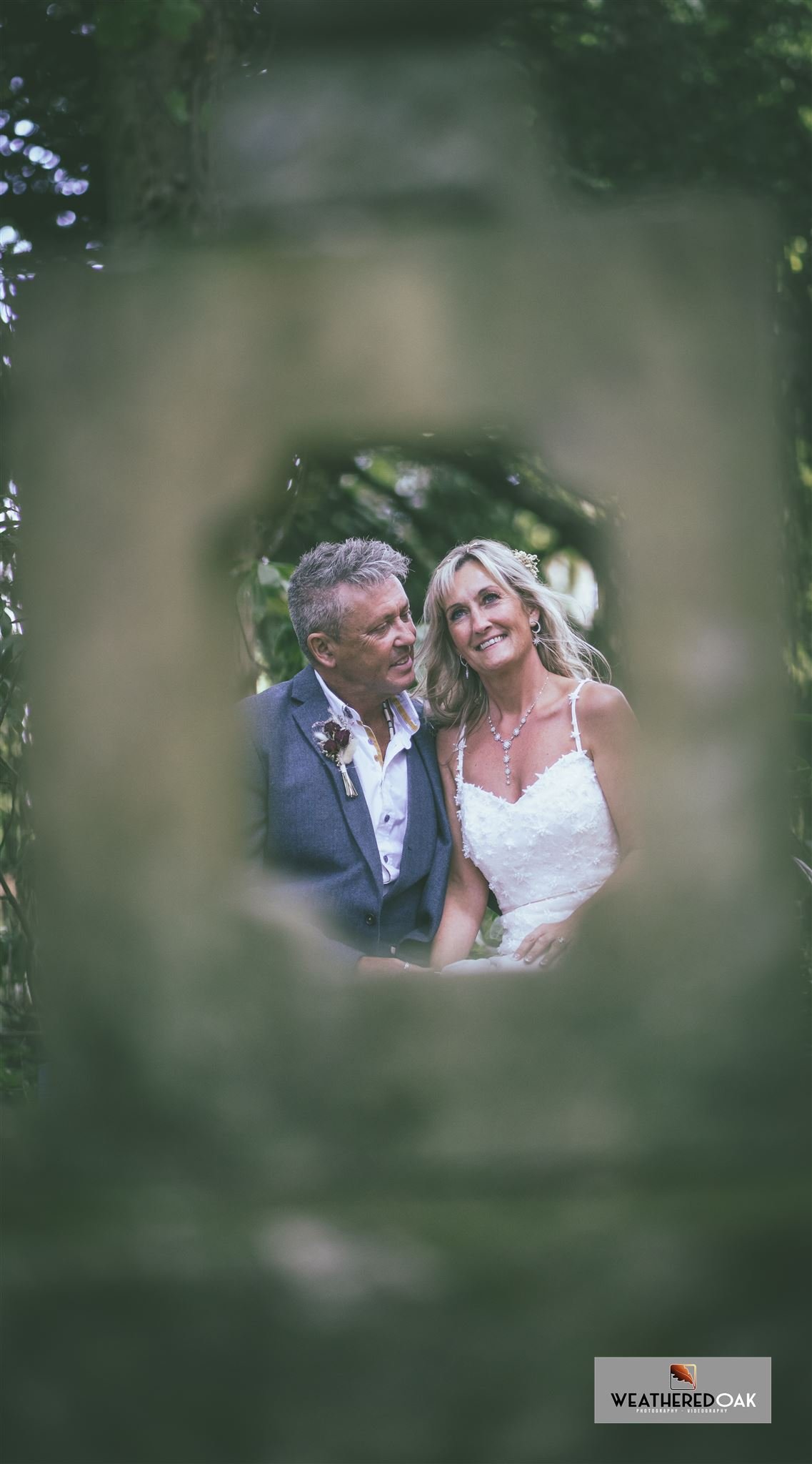 A wedding couple is seen through a blurred frame, with the bride in a white dress and the groom in a suit, set outdoors with greenery in the background.