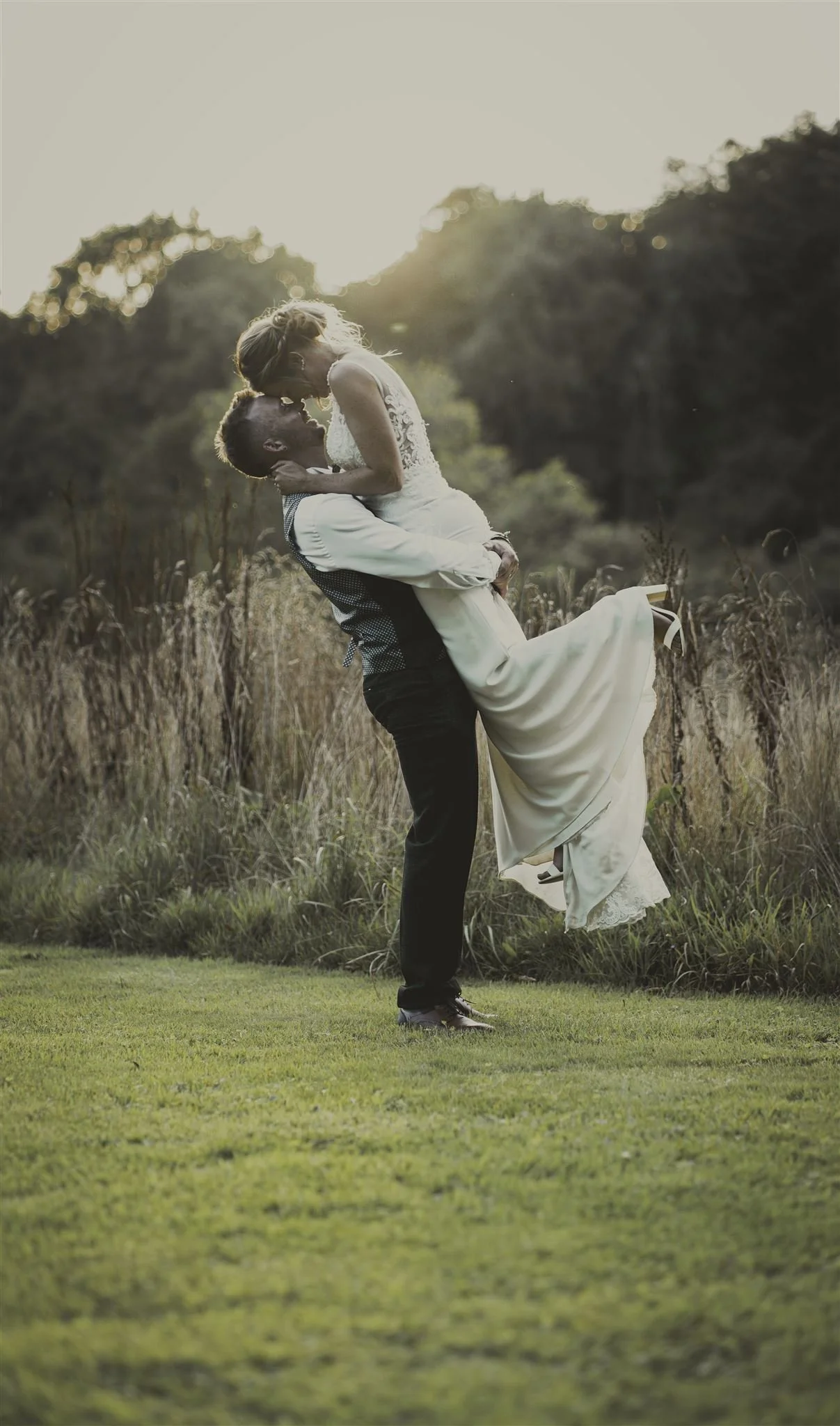 A couple in wedding attire embracing in a grassy field at sunset; the groom lifting the bride in his arms.