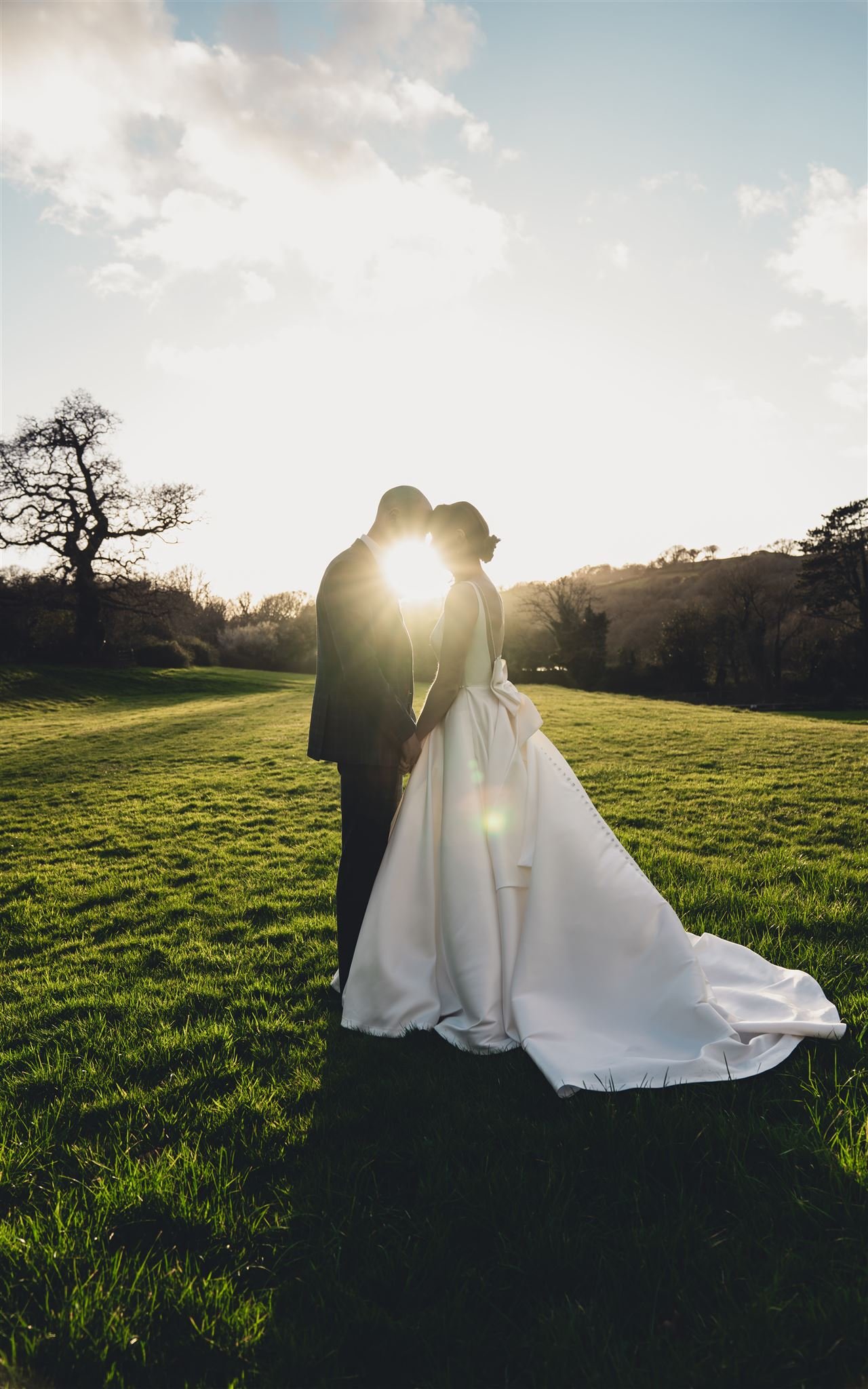 Bride and groom standing in a field at sunset, holding hands and facing each other, with the sun shining between them.