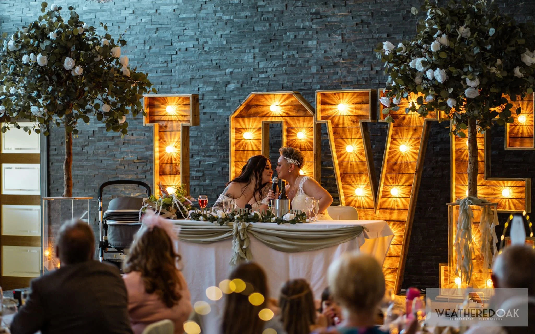 Wedding reception scene with two women at a decorated table, large illuminated "LOVE" letters in the background, and guests seated in the foreground. The setting is elegant with floral arrangements and dim lighting.