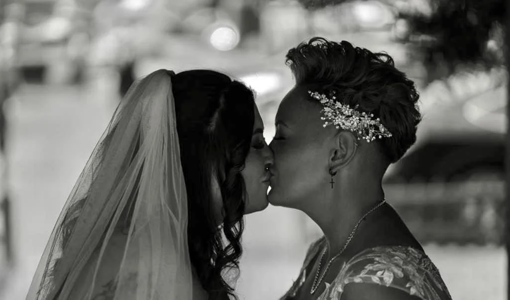 Two women kissing at a wedding, one wearing a veil and the other with short hair and a decorative headpiece.