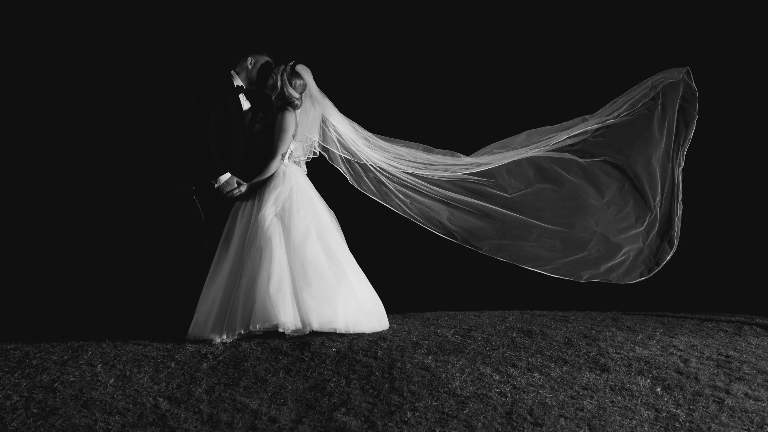 Black and white photo of a bride and groom kissing, bride's veil flowing, nighttime setting.