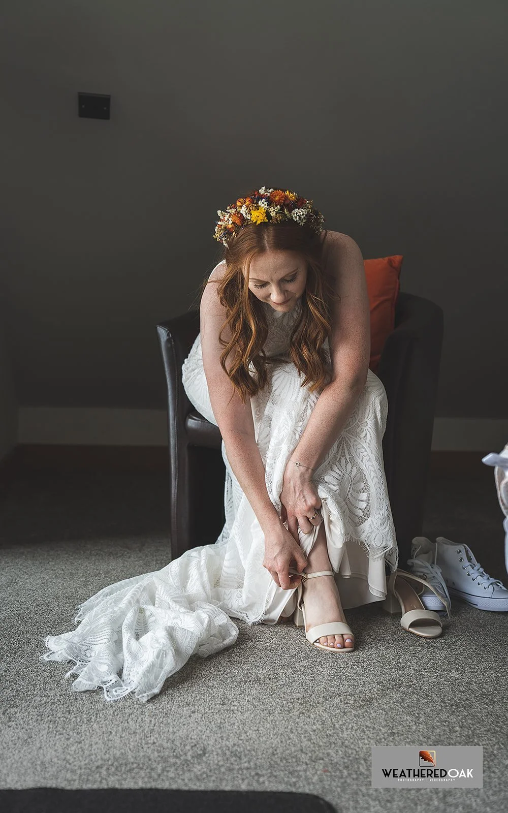 Bride wearing a floral crown and wedding dress adjusts her sandal strap while sitting in a chair, with sneakers on the floor nearby.