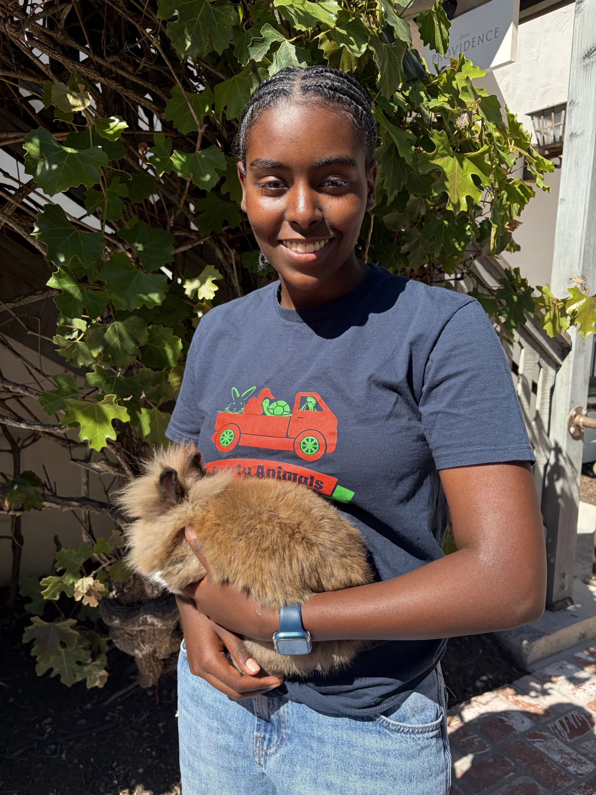 A young woman smiling and holding a fluffy, orange rabbit outdoors with green ivy and plants in the background.