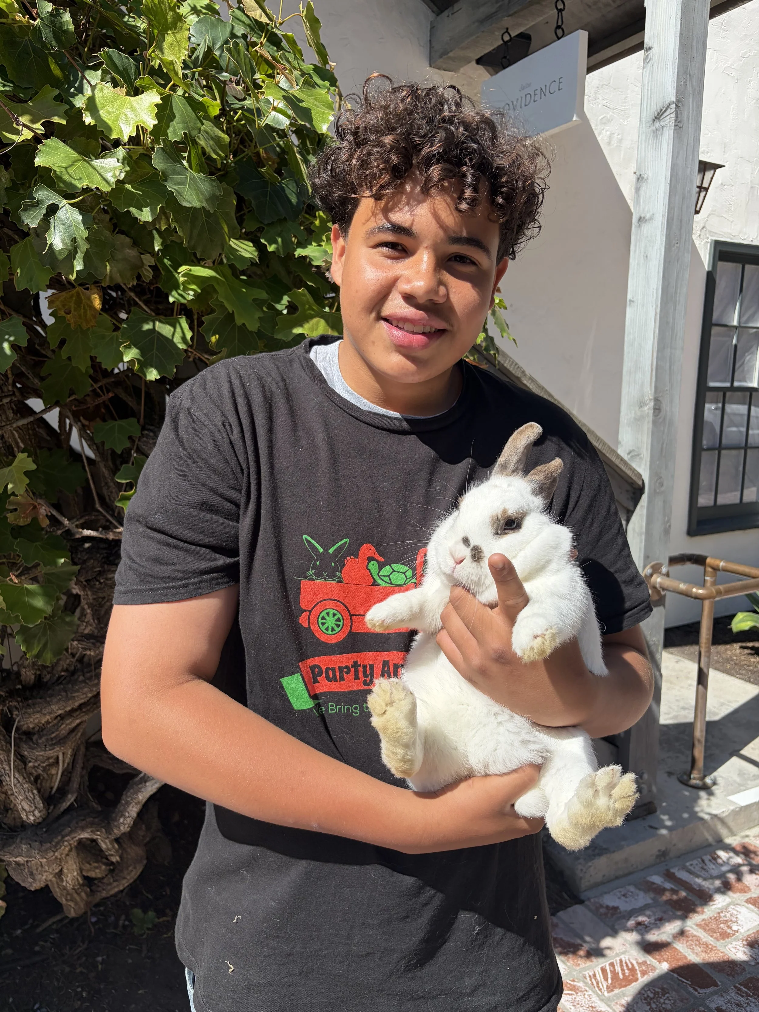 A young man with curly brown hair holding a white bunny rabbit with black markings outdoors on a sunny day, standing beside a leafy green hedge and a white building.