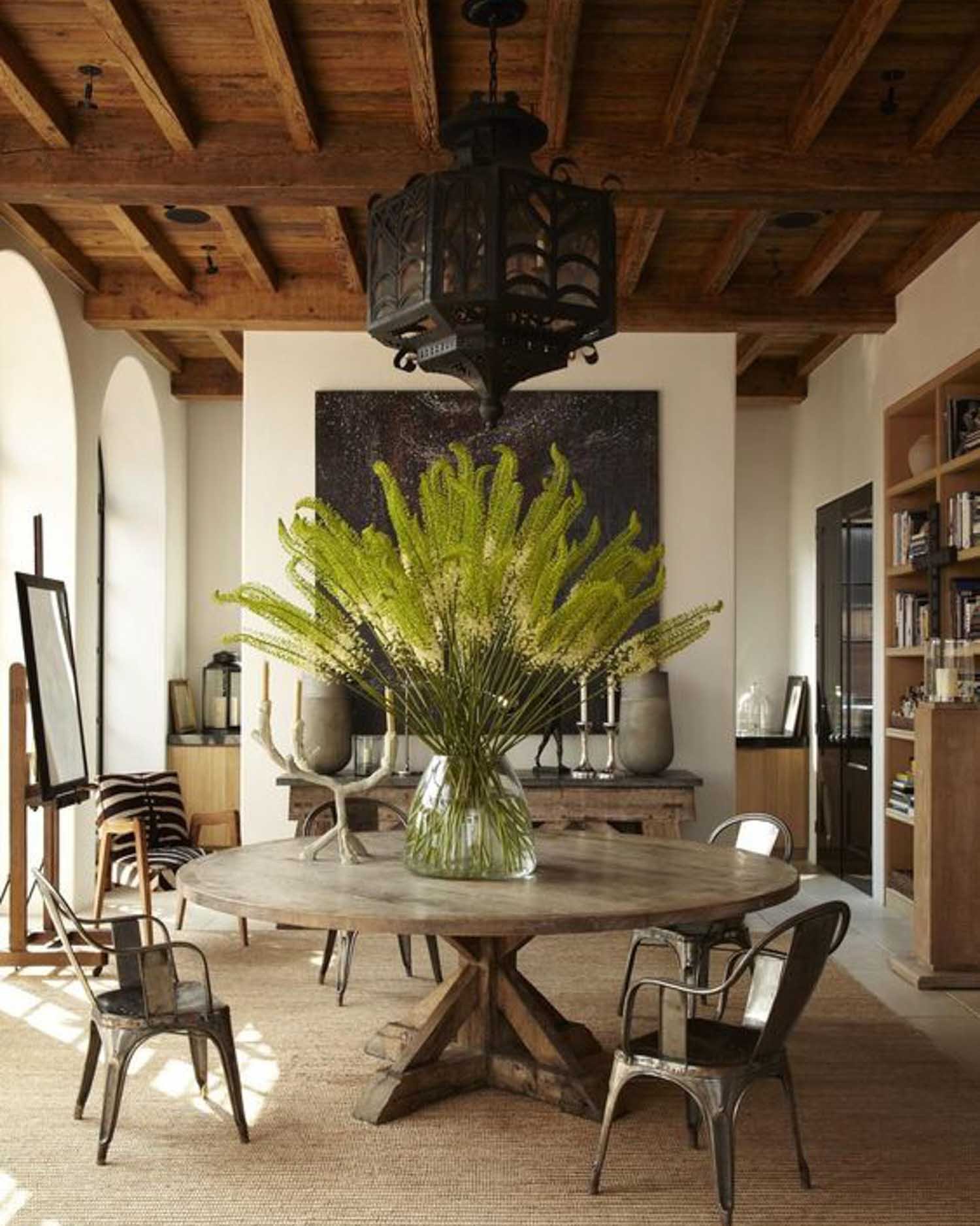 A large display of fox tail lilies in a glass vase sit on a round wooden table in a dining room with a wooden vaulted ceiling and arched glass doors