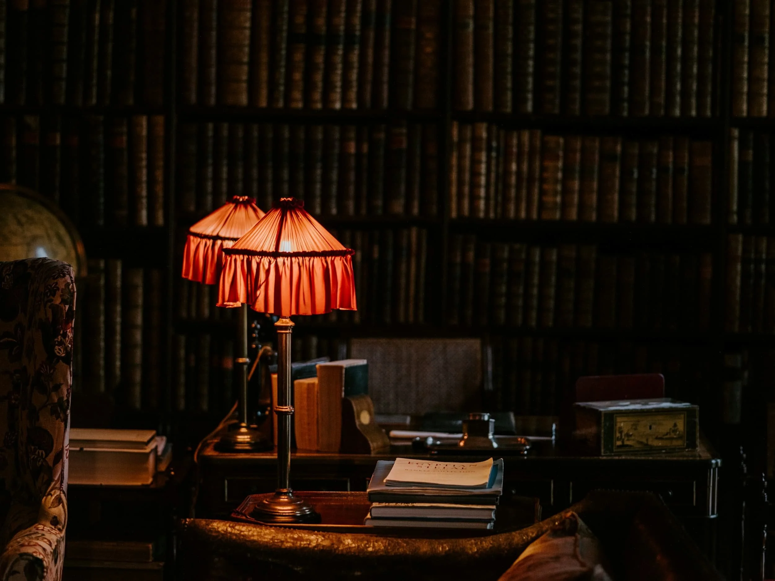 Two table lamps with red cloth shades in a dark library next to an armchair with cracked leather and full bookshelves behind